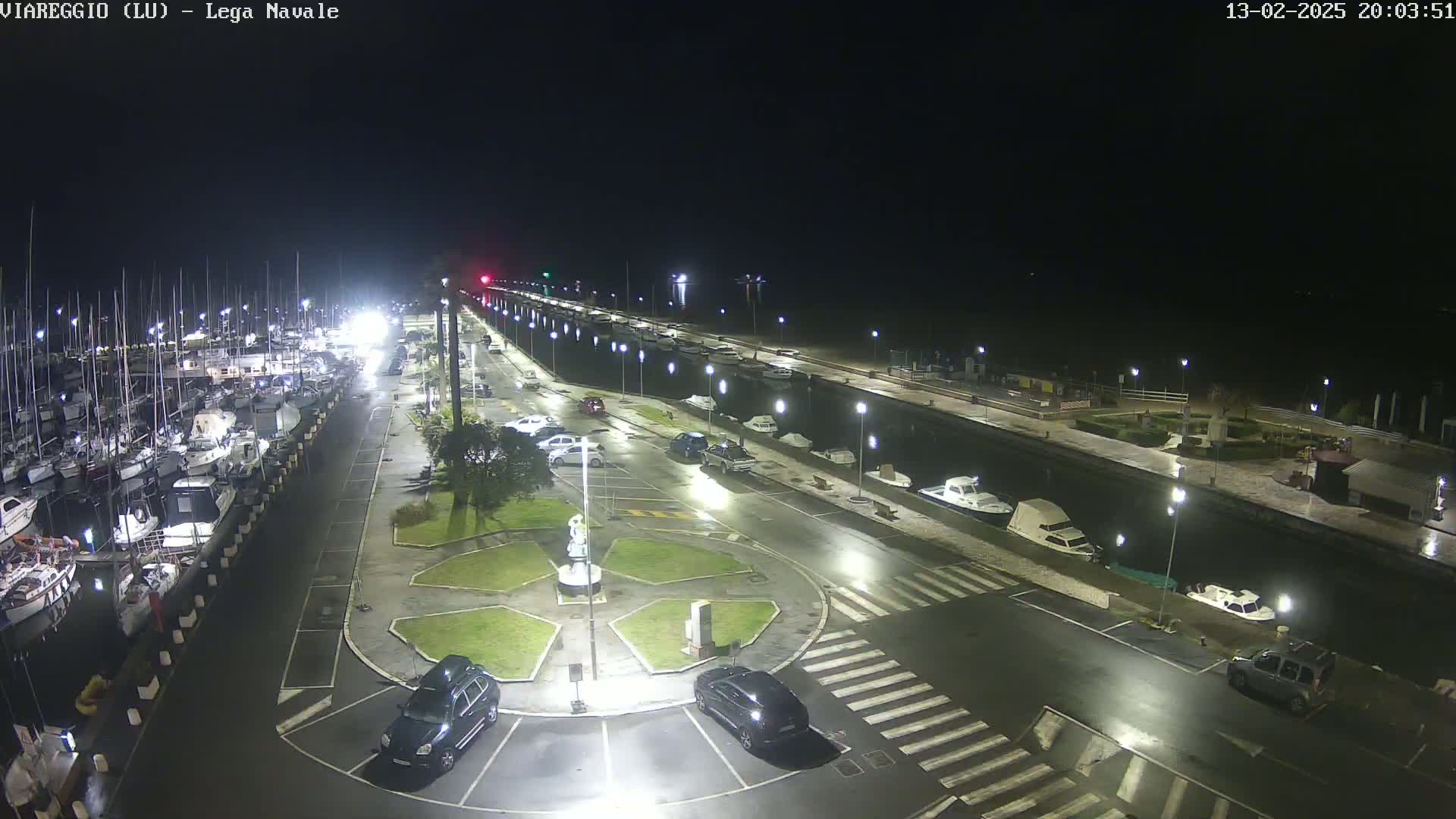 A nighttime, wet marina scene shows numerous boats docked along a waterfront, with cars parked in lots lining a canal, and a small roundabout in the foreground.