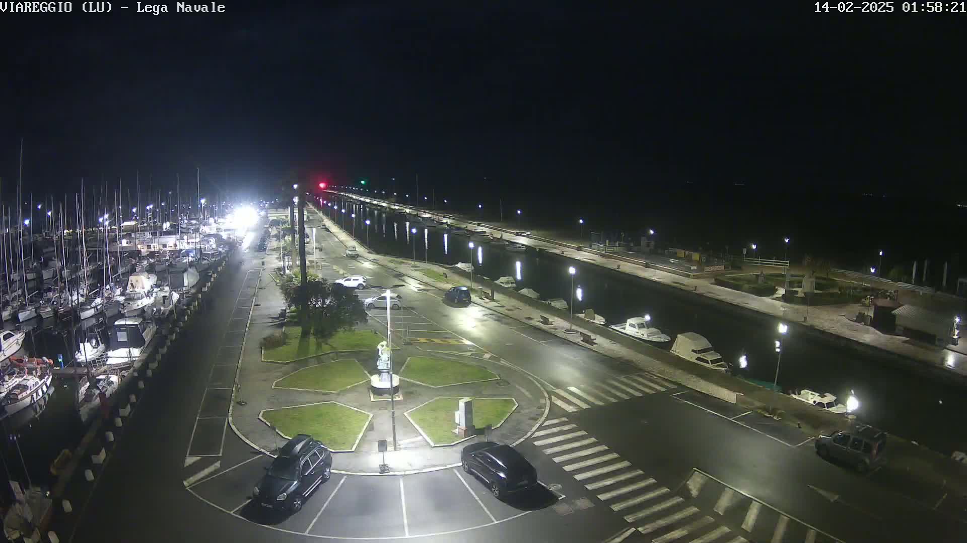 A nighttime view of a marina with docked boats, cars parked in a lot, and a small roundabout in the center, under slightly wet conditions.