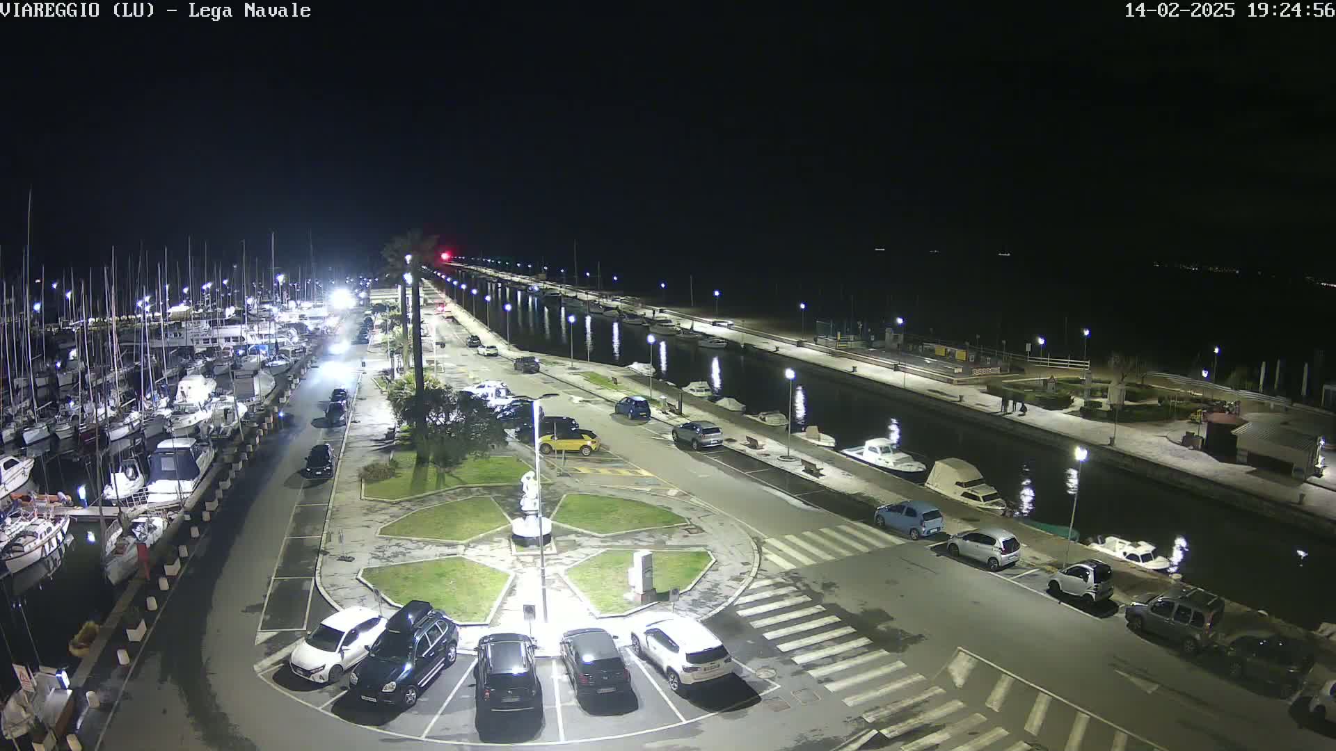 A nighttime aerial view of a marina with numerous boats docked, a paved area with parked cars, and a central plaza featuring a statue, all illuminated by artificial lighting.