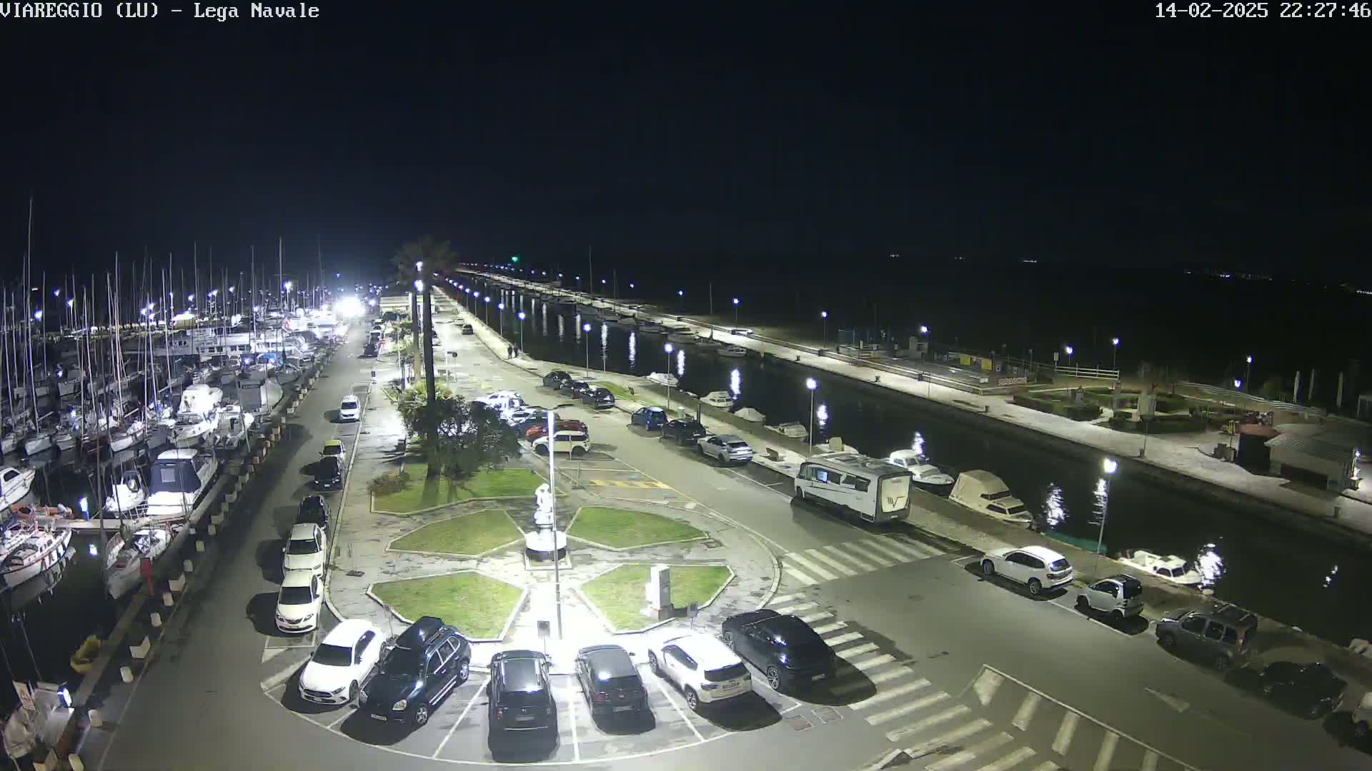 A nighttime aerial view shows a marina filled with boats, a paved area with parked cars surrounding a central landscaped area, and a canal reflecting the lights.