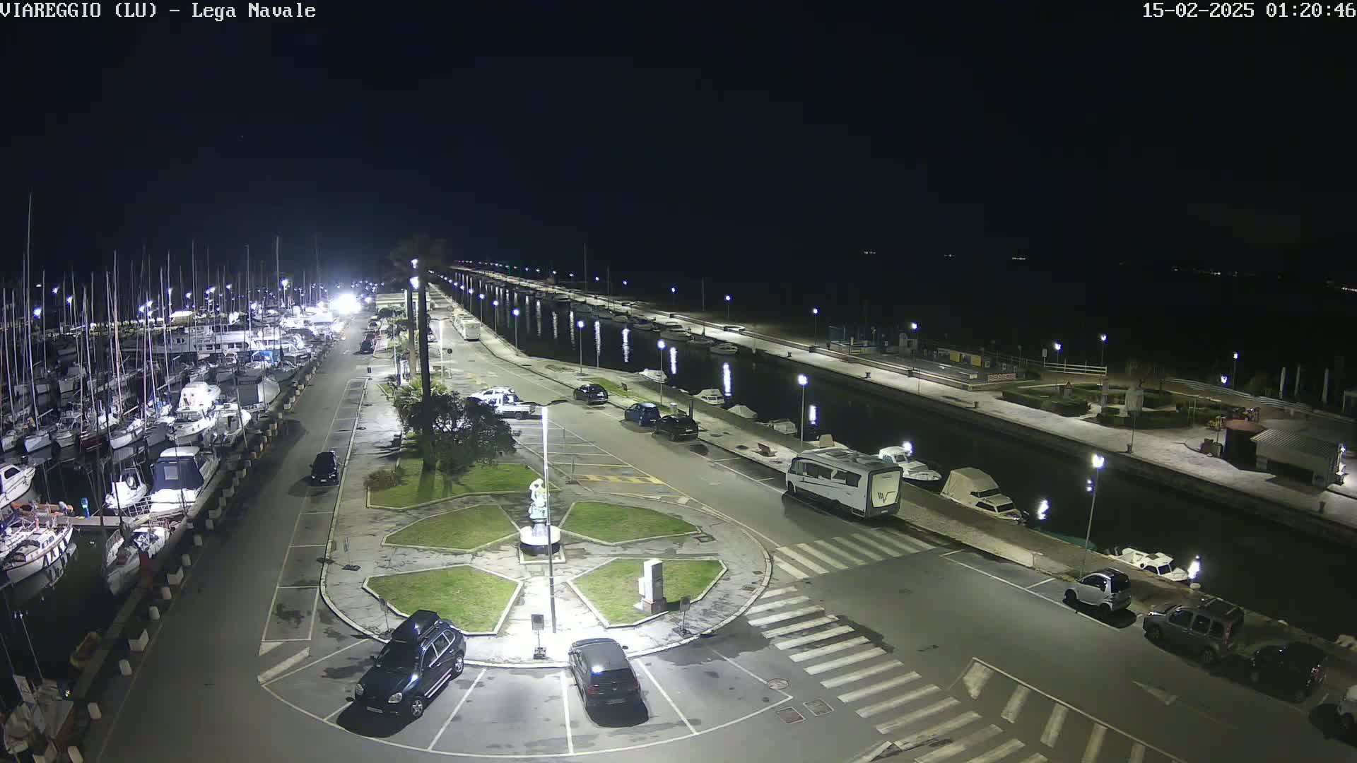 A nighttime aerial view of a marina with numerous boats docked alongside a paved area with cars parked, a central roundabout, and streetlights illuminating the scene.