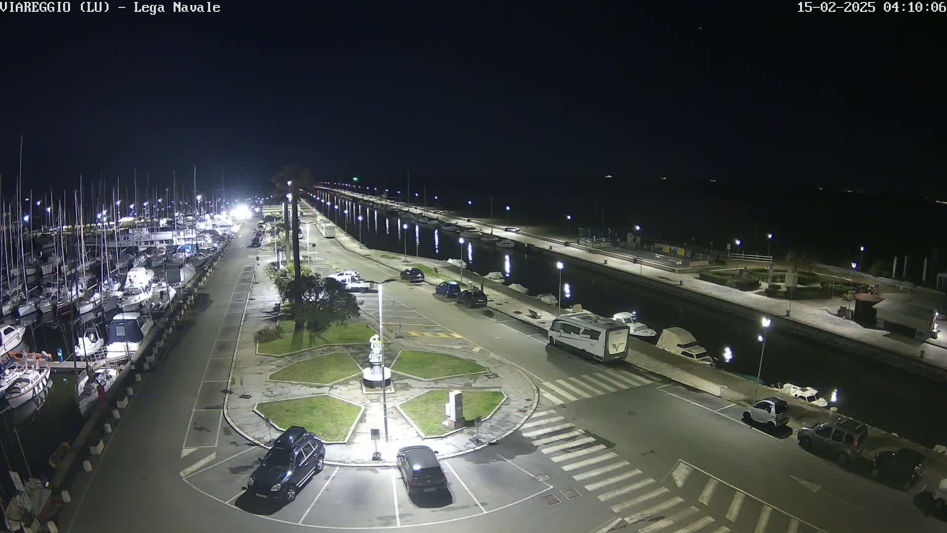 A nighttime aerial view of a marina with numerous boats docked, a roadway with parked cars, and a central roundabout, all brightly lit under a clear, dark sky.