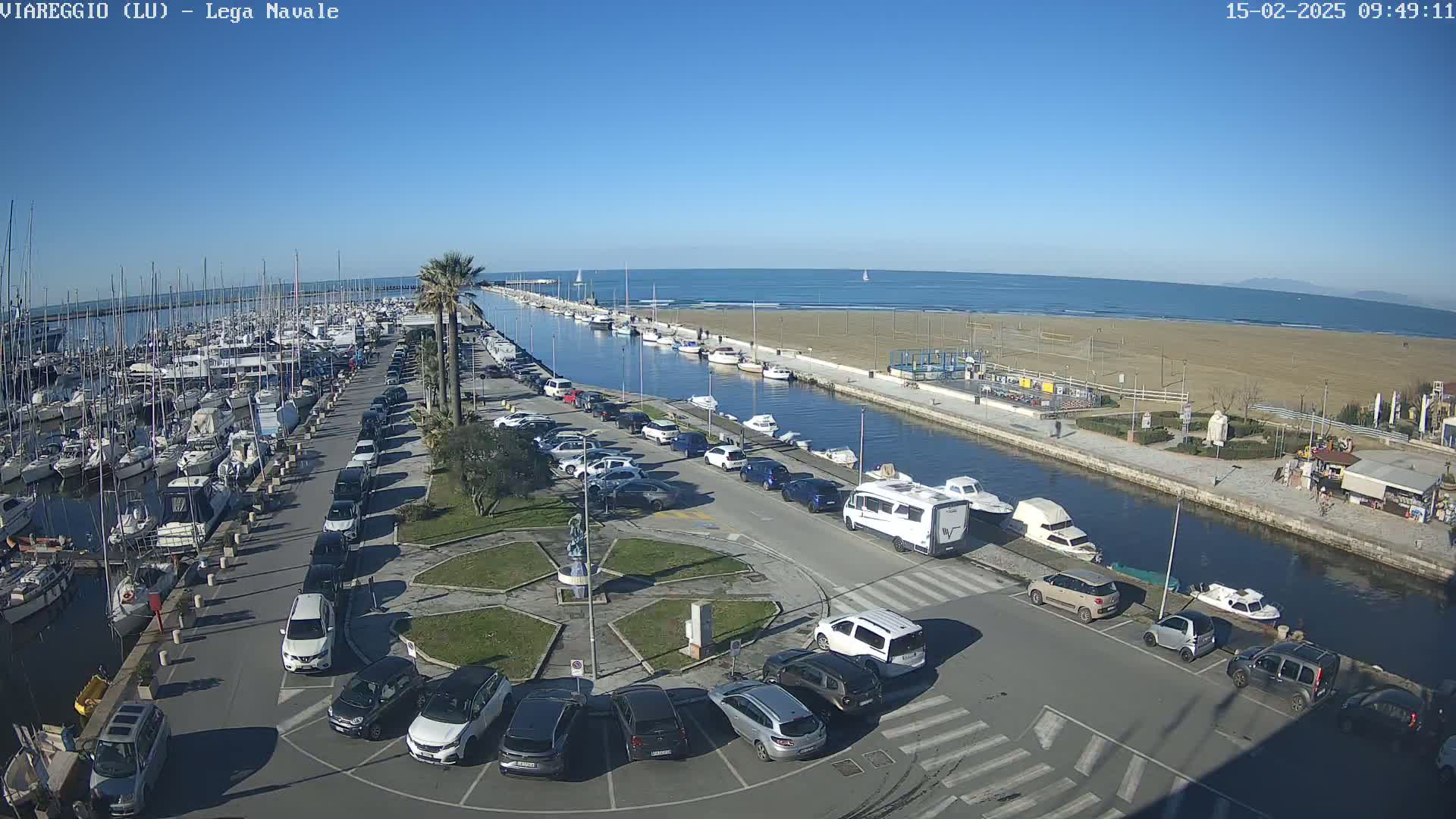 A marina filled with boats is adjacent to a sandy beach and a canal lined with parked cars under a clear blue sky.