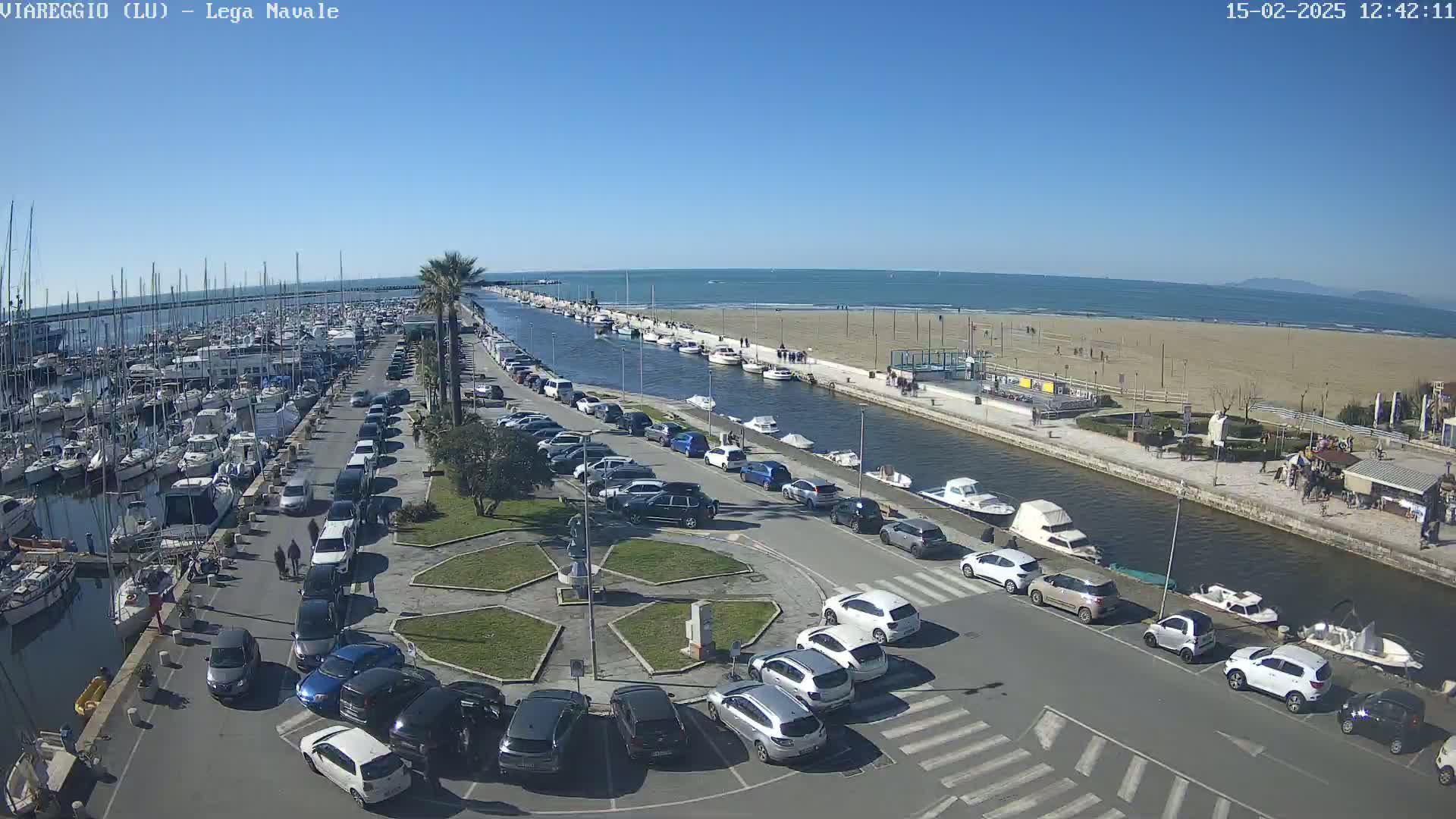 A marina filled with boats is adjacent to a sandy beach and calm ocean under a clear blue sky.