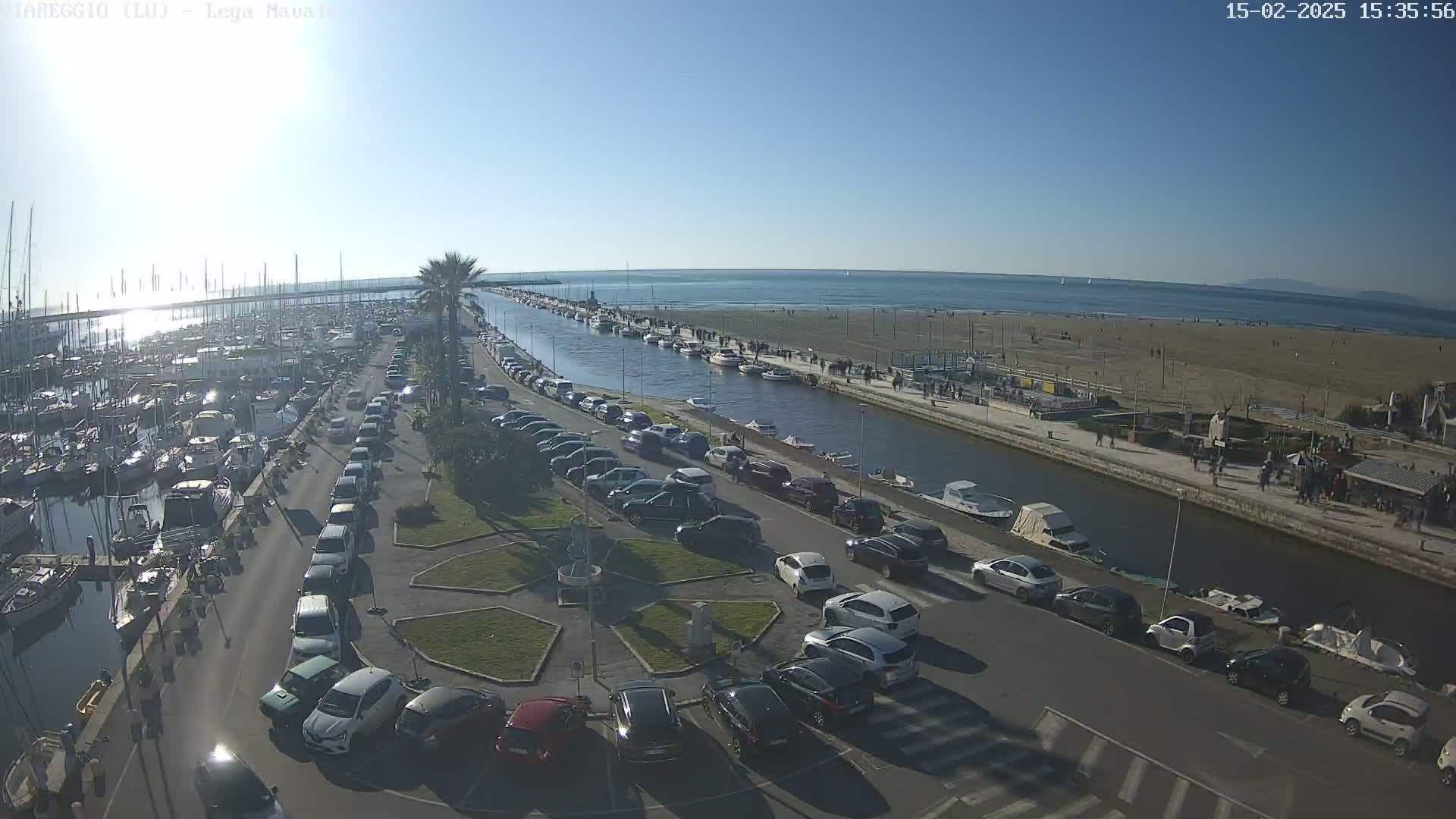 A marina filled with boats is next to a canal, which runs alongside a sandy beach with people walking along the shore under a clear, sunny sky.