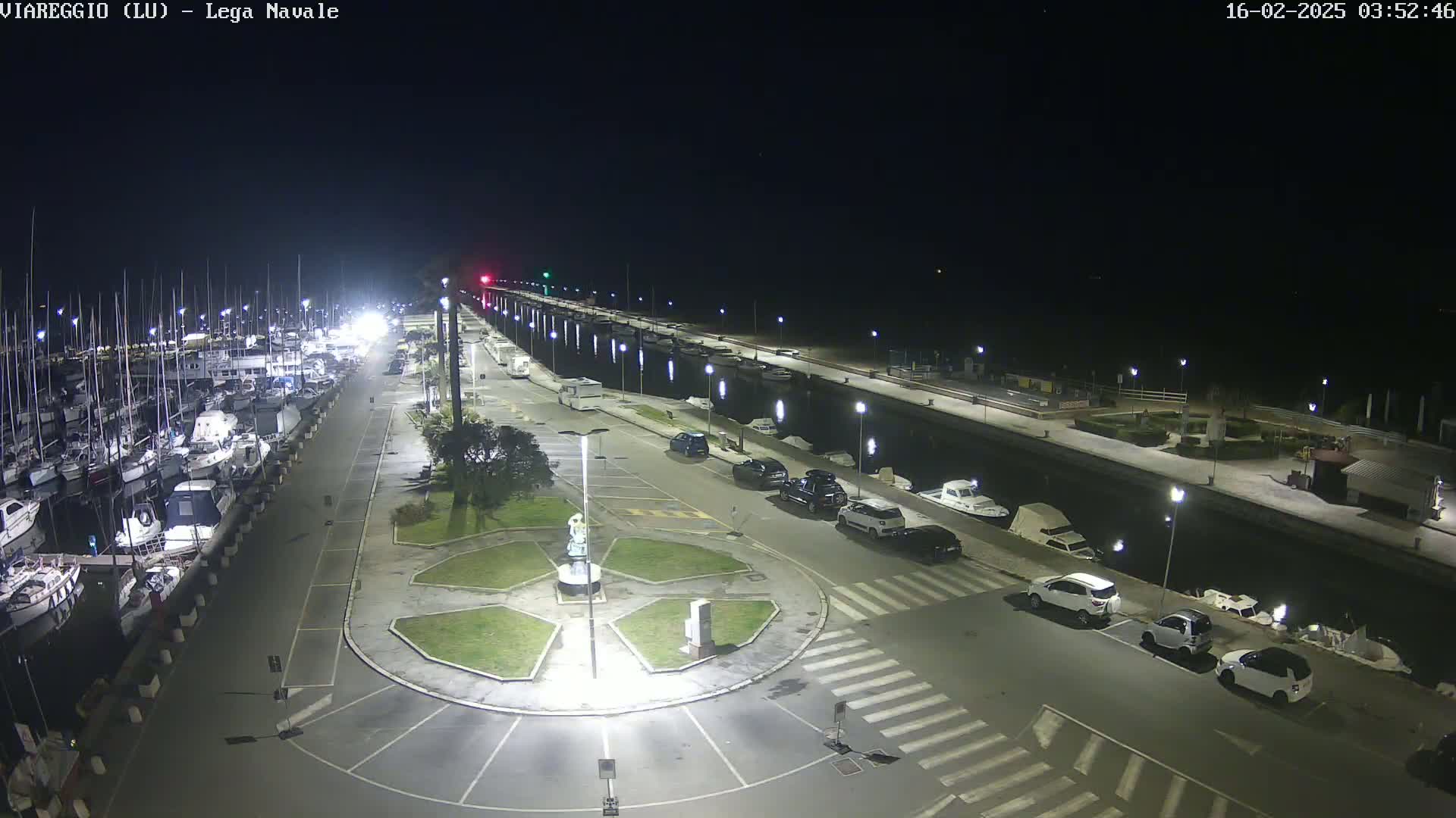 A nighttime, brightly lit view of a marina filled with boats, a roadway with parked cars, and a small circular plaza with a statue in the center.
