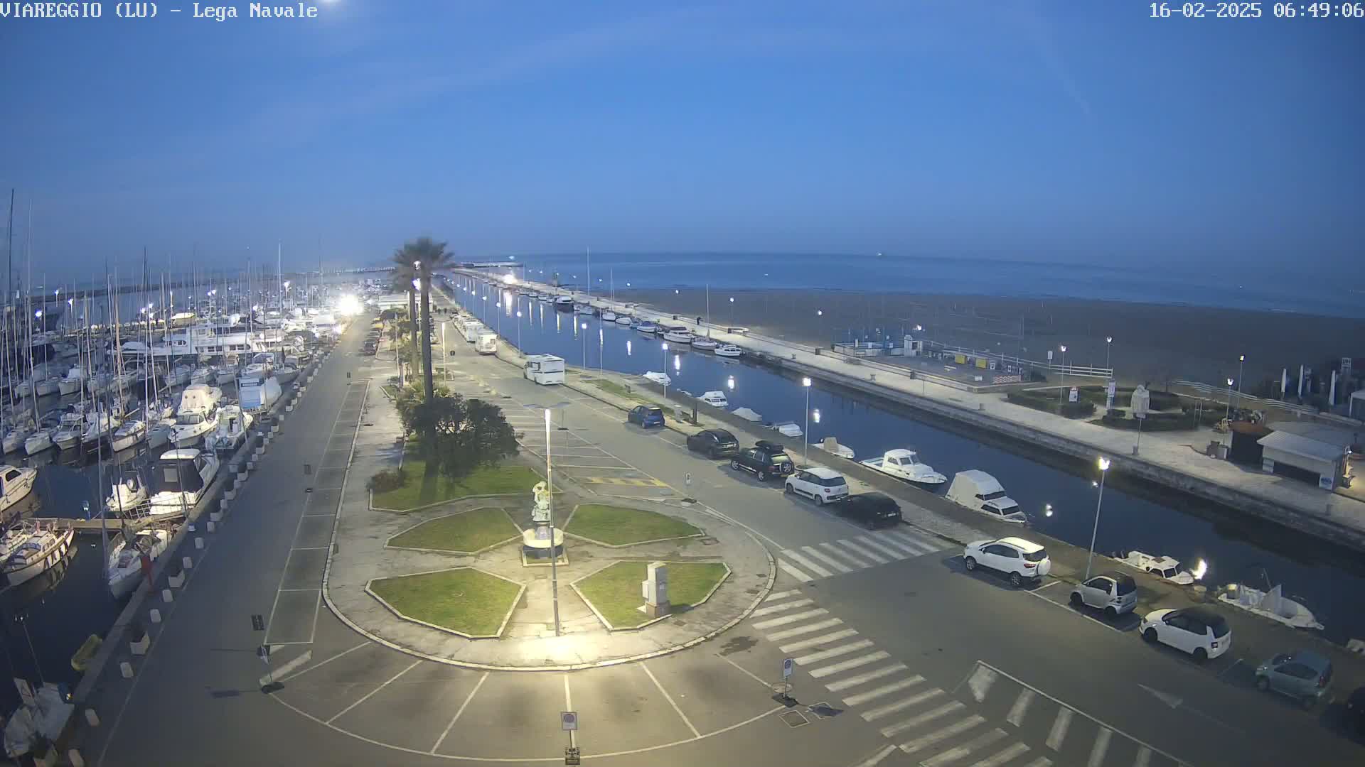 A marina filled with boats is adjacent to a calm, dark-blue ocean under a twilight sky, with a paved area containing several parked cars and a small plaza in between.