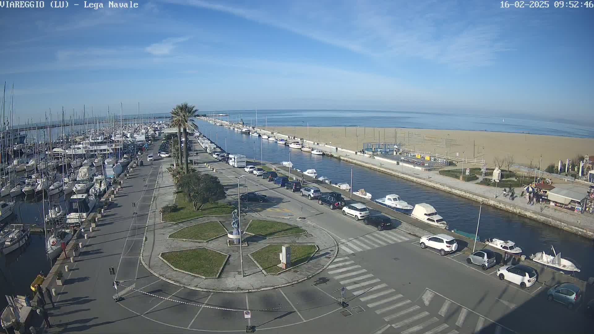 A marina filled with boats is adjacent to a canal and a sandy beach under a clear blue sky.