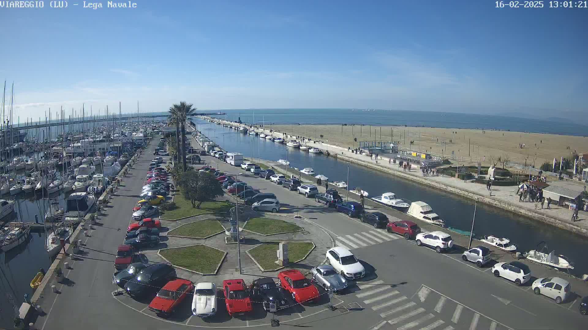 A marina filled with boats, a canal lined with parked cars and pedestrians, and a sandy beach under a clear blue sky.