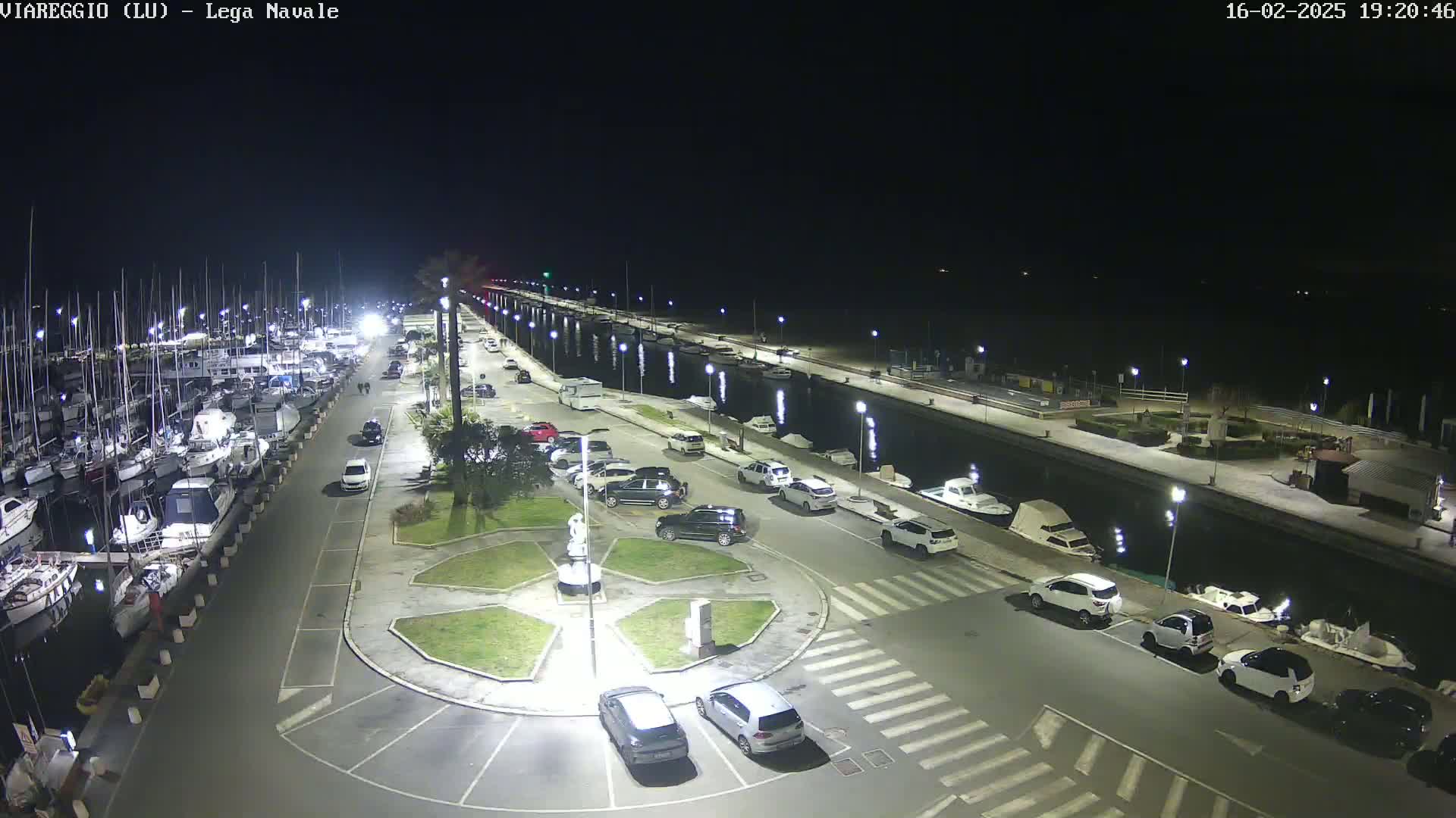 A nighttime aerial view of a marina with numerous boats docked, a parking lot filled with cars, and a central roundabout, all brightly lit.