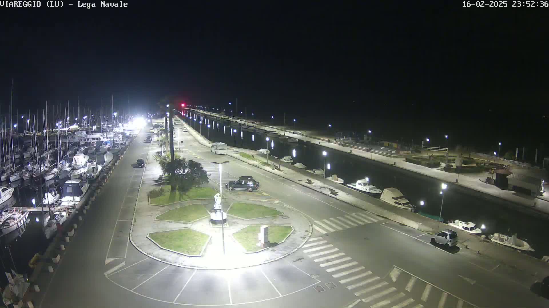 A nighttime aerial view of a marina with numerous boats docked, a roadway with cars parked, and a central roundabout with a statue, all brightly lit under a clear, dark sky.