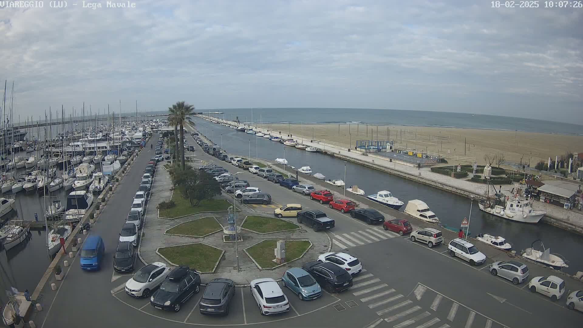 A marina filled with boats is next to a canal, which leads to a sandy beach and calm ocean under a cloudy sky.  Cars are parked along the canal and in a nearby parking lot.