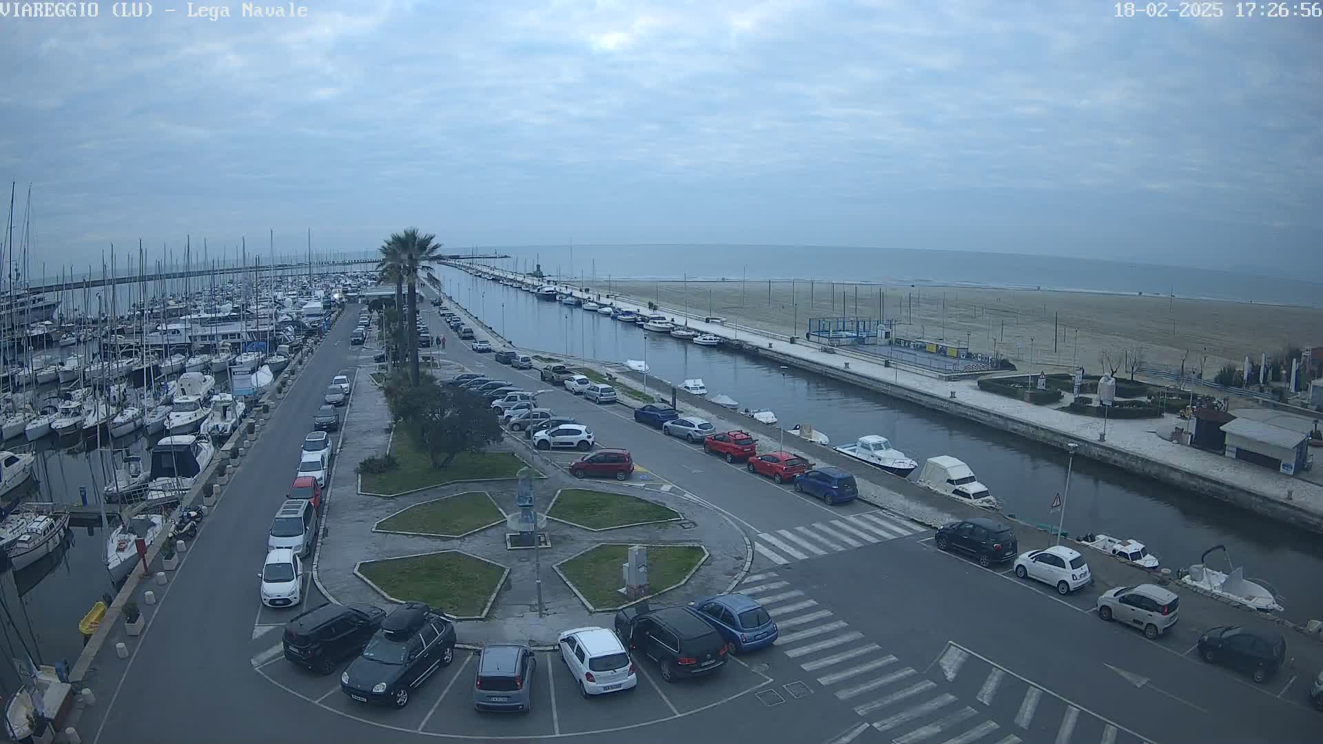 An overcast day shows a marina filled with boats, a canal lined with parked cars, and a beach in the distance.