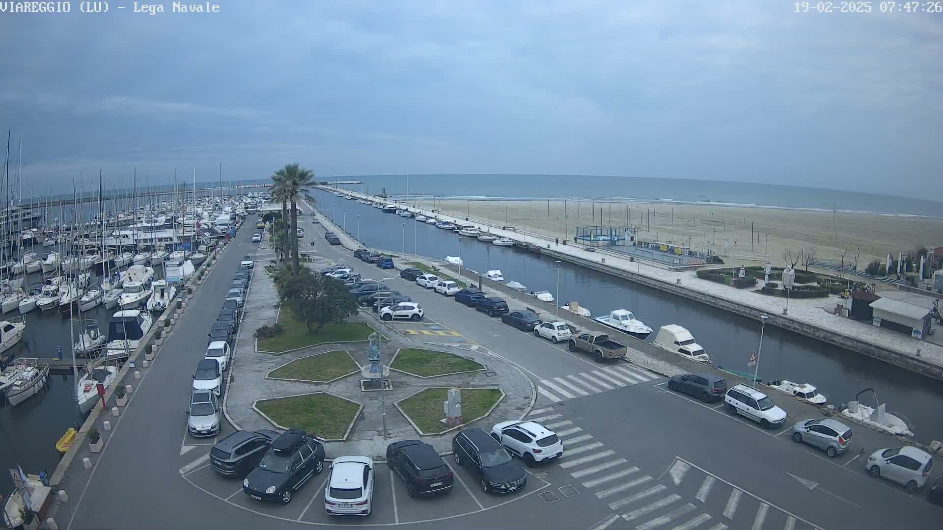A marina filled with boats is adjacent to a parking lot and canal leading to a sandy beach under a cloudy sky.