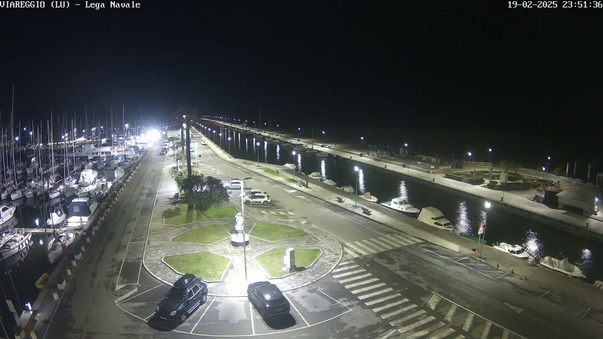 A nighttime aerial view of a marina with docked boats, cars parked in a lot, and a canal reflecting lights.