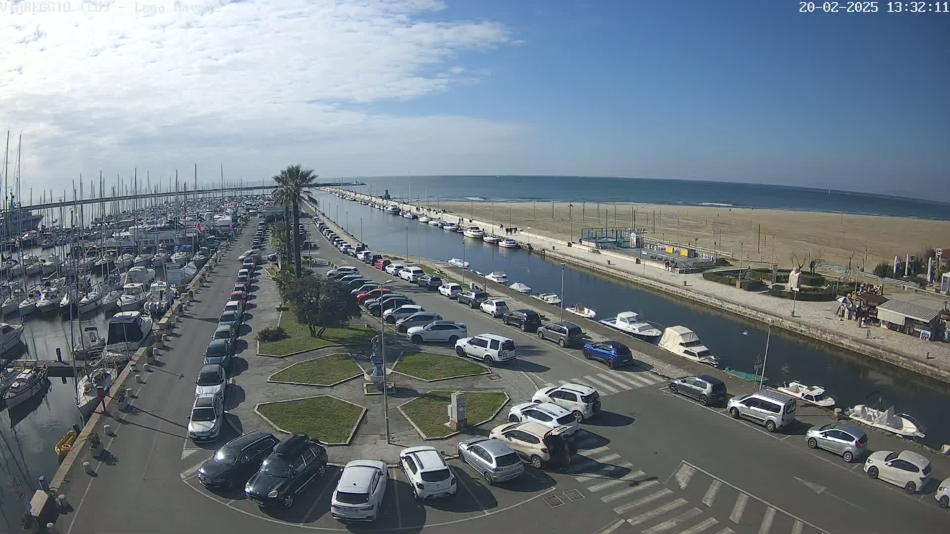 A marina filled with boats and surrounded by a parking lot overlooks a calm ocean on a sunny day.