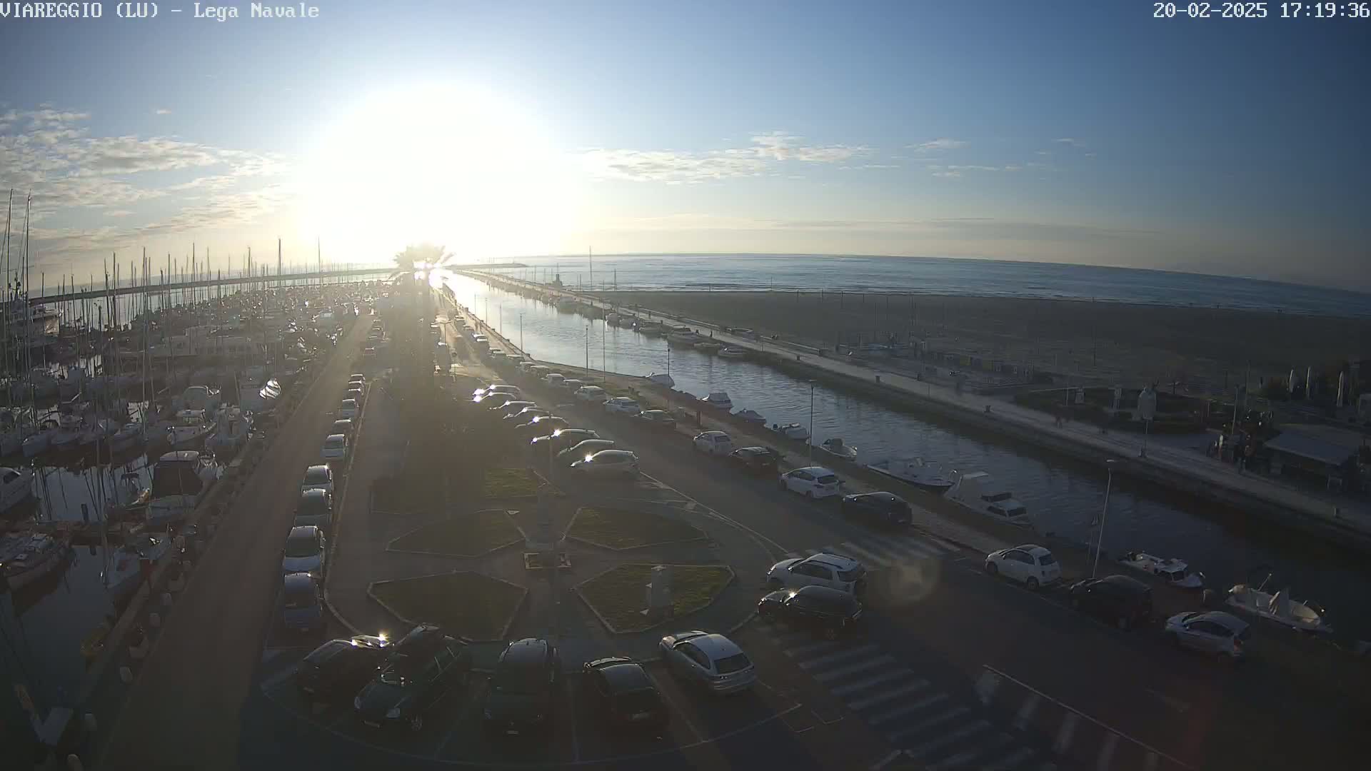 A marina filled with boats is adjacent to a canal and beach under a mostly clear sky, with the sun setting over the ocean in the distance.