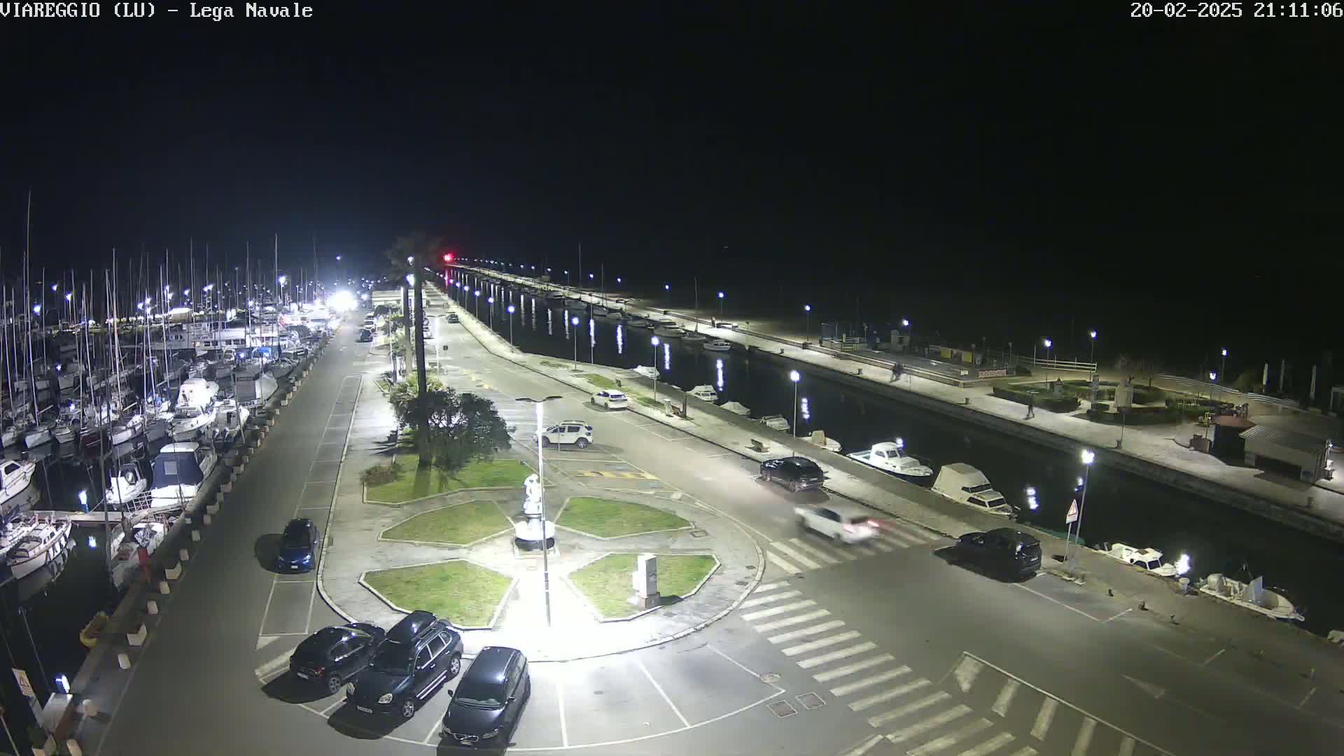 A nighttime view of a marina with numerous boats docked along a canal, several cars parked in a nearby lot, and a statue in a circular plaza.
