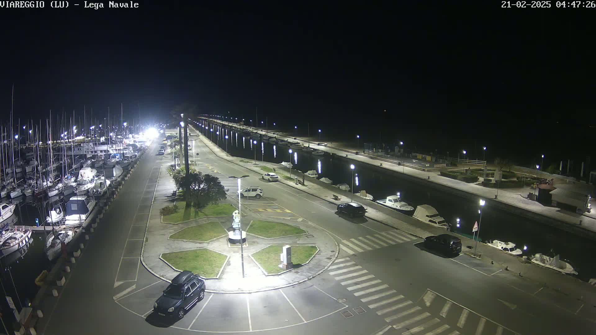 A nighttime, brightly lit marina with numerous boats docked alongside a paved area containing several parked cars and a central roundabout.