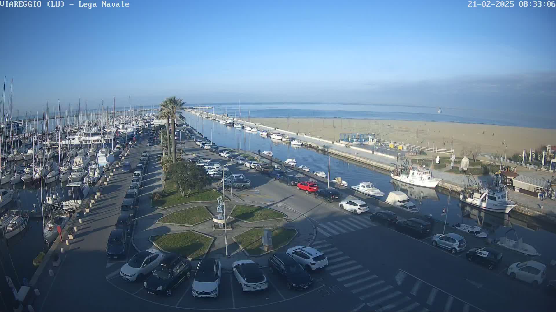 A marina filled with boats, a sandy beach, and a parking lot with cars are visible on a clear, sunny day.
