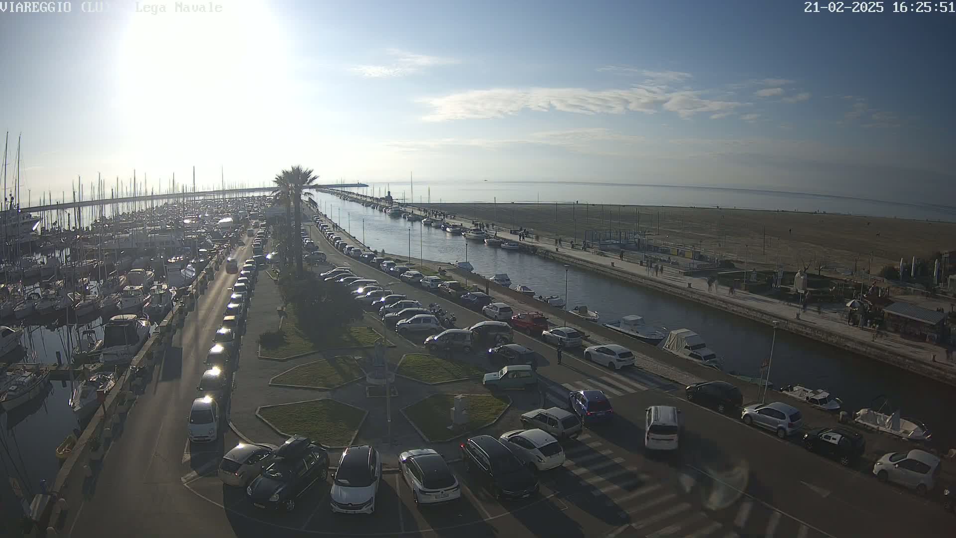 A marina filled with boats and surrounded by parked cars, next to a calm body of water under a clear, sunny sky.