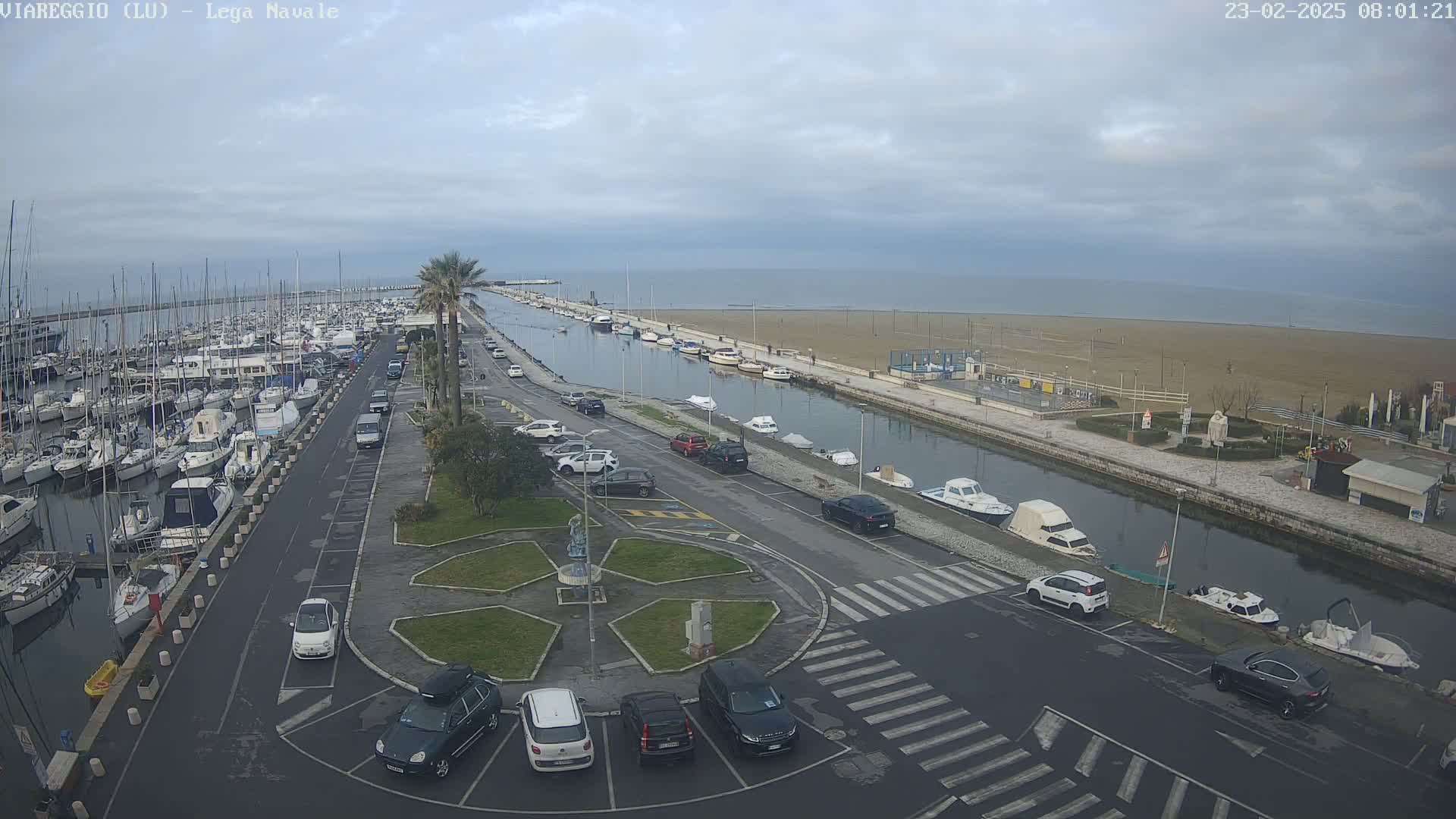 An overcast day shows a marina filled with boats, a canal with additional boats, a sandy beach, and a parking lot with cars next to a roundabout.