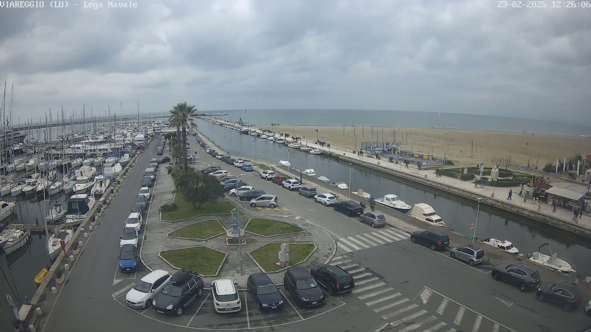 A marina filled with boats is next to a canal lined with parked cars and leading to a sandy beach under an overcast sky.