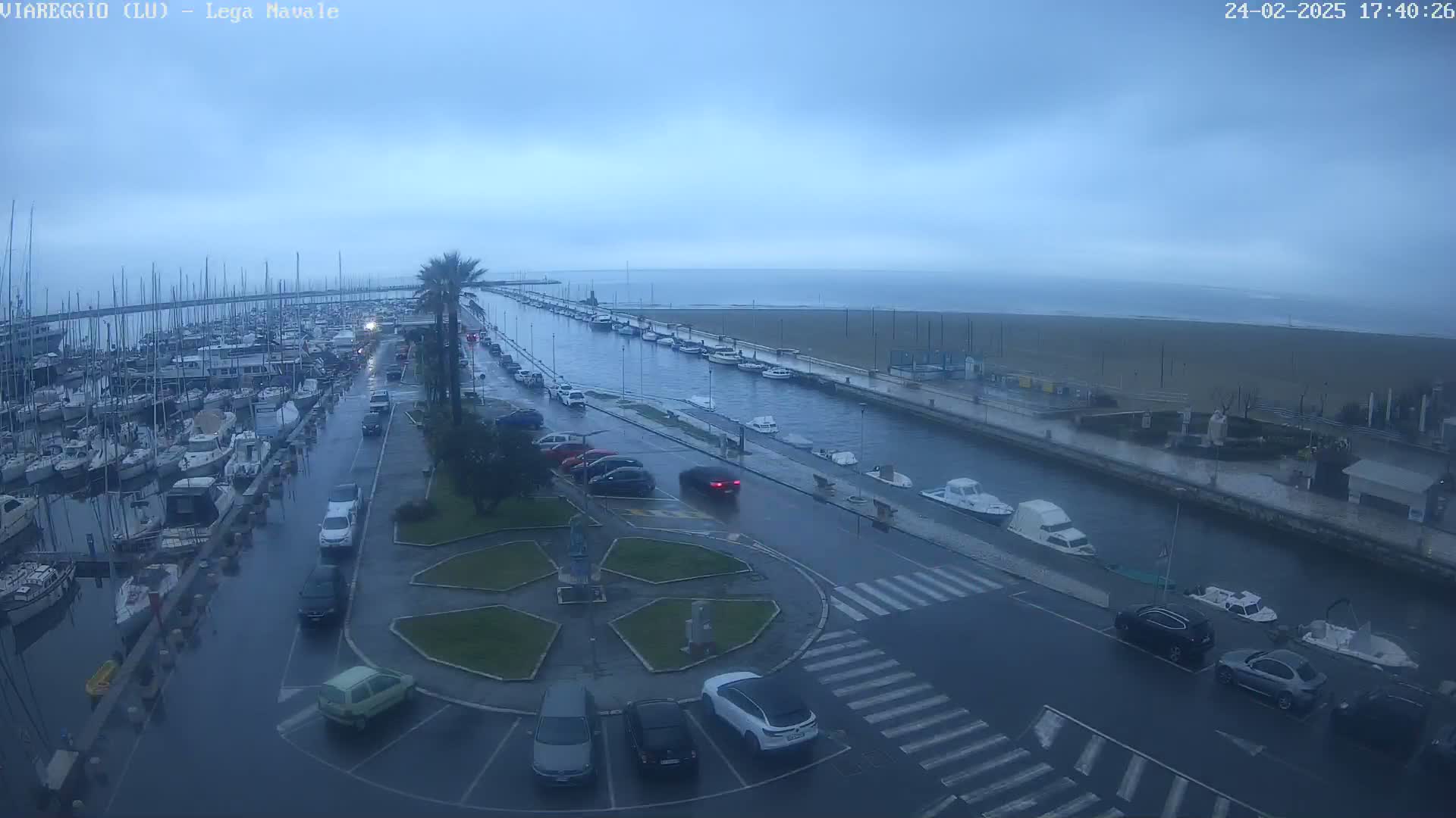 A wet, overcast day reveals a marina filled with boats, a canal with moored vessels, and a roadway with parked cars, all overlooking a beach under a gray sky.