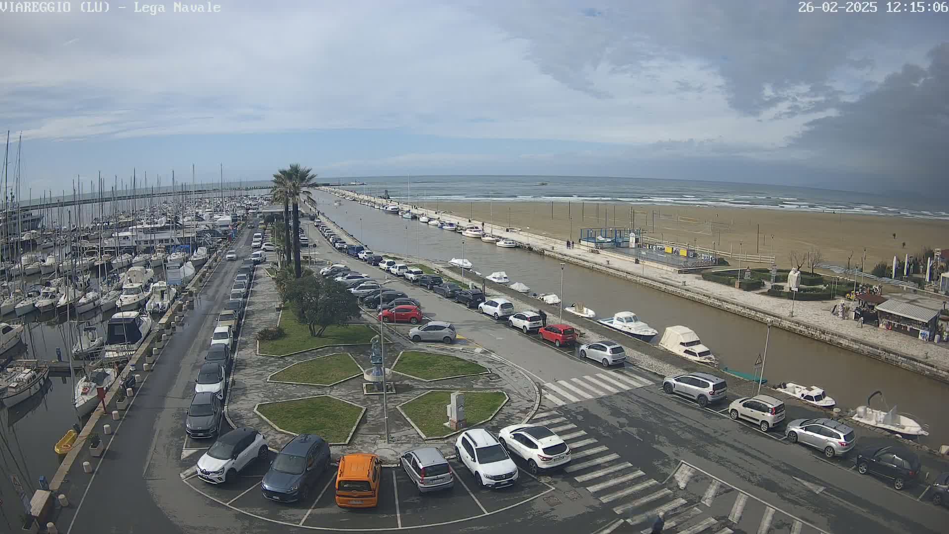 A harbor filled with boats and surrounded by parked cars is next to a sandy beach with a calm ocean under a mostly cloudy sky.