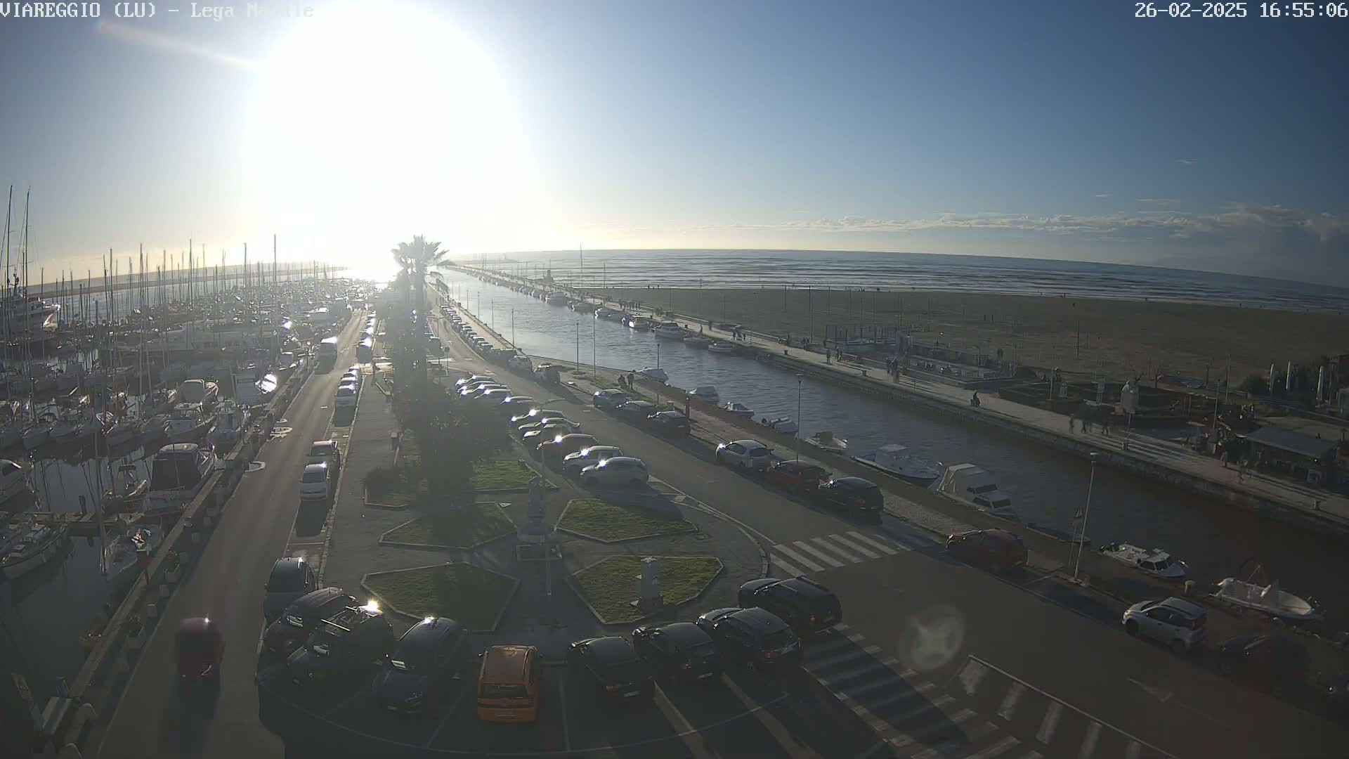 A sunny day shows a marina filled with boats, a canal, a beach, and cars parked along a road, all under a mostly clear sky.