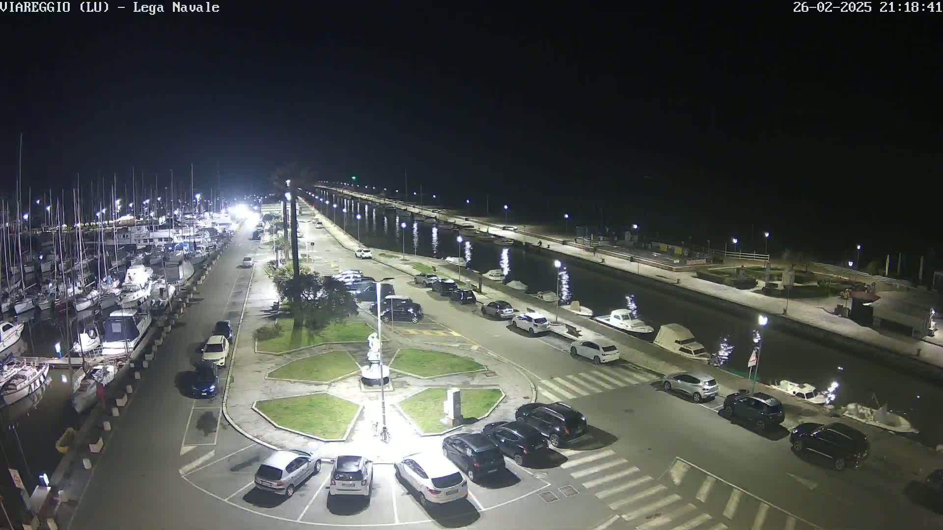 A nighttime aerial view of a marina with numerous boats docked, a canal, and a parking lot filled with cars surrounding a small, landscaped central area.