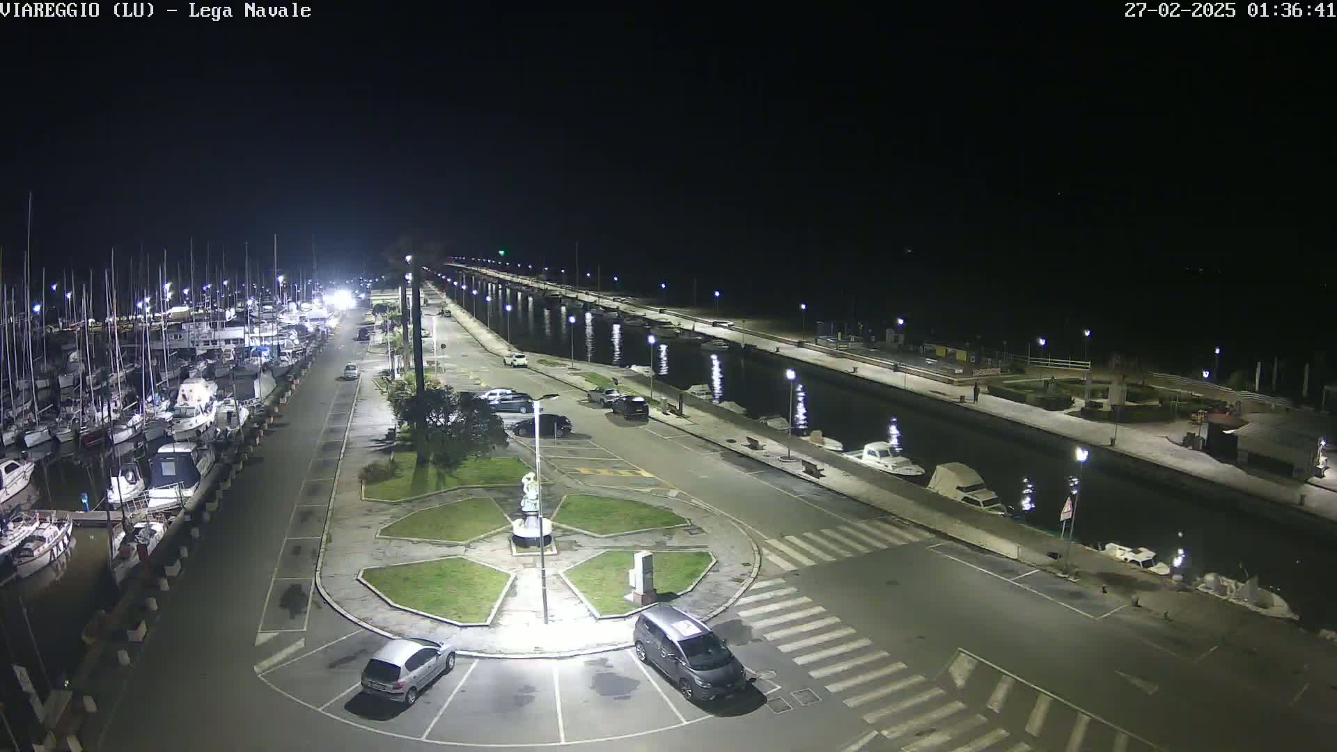 A nighttime view of a marina with numerous boats docked, a calm canal, and a paved area with parked cars and a small roundabout.