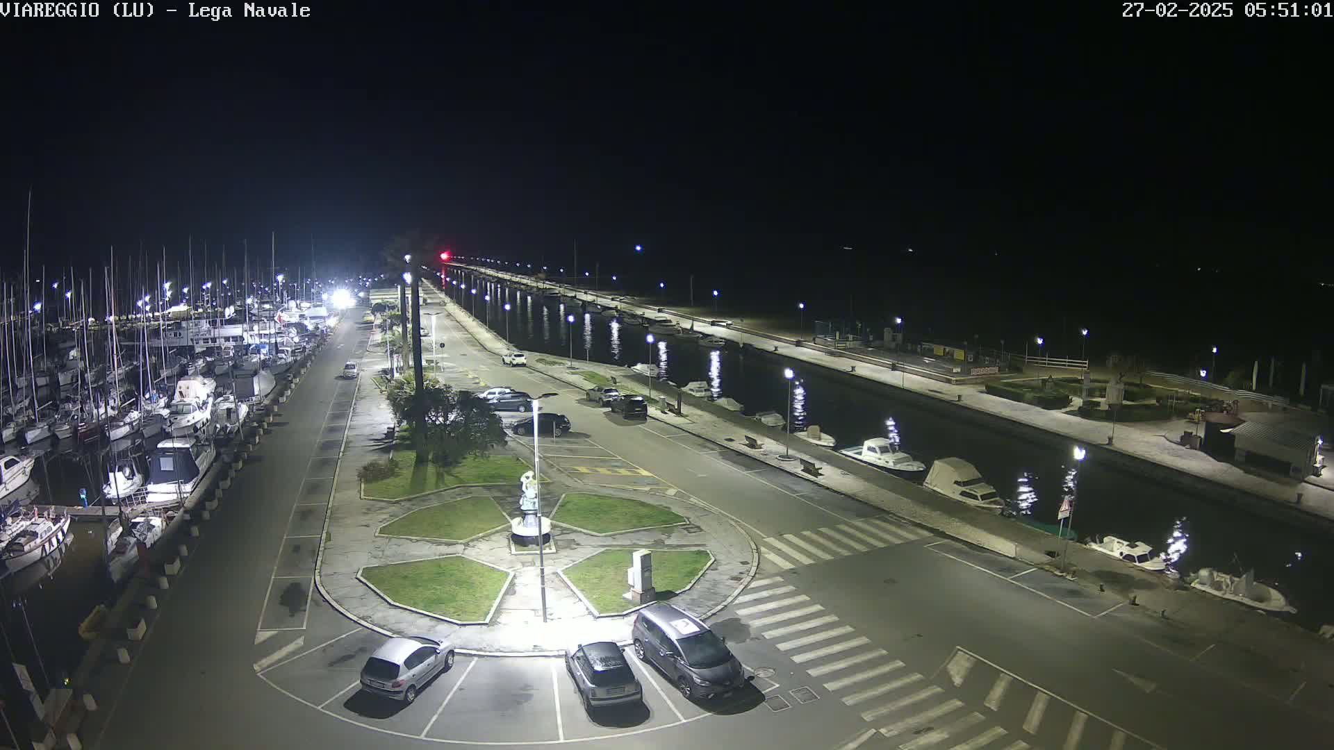 A nighttime view of a marina with numerous boats docked along a waterfront, a roadway with parked cars, and a central roundabout.