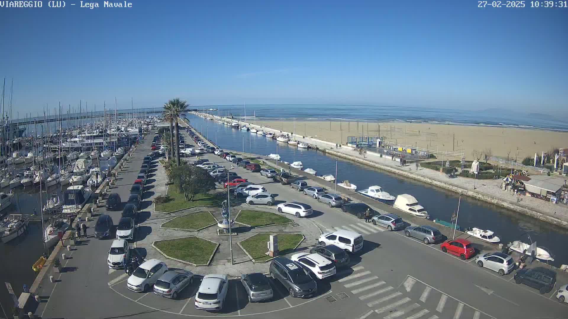 A sunny day overlooking a marina filled with boats, a canal, a sandy beach, and a parking lot full of cars.