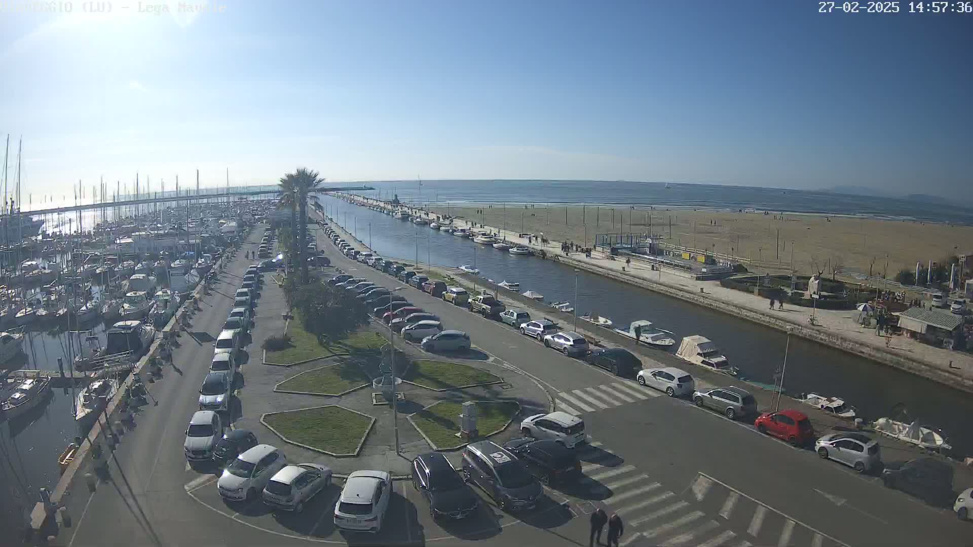 A sunny day at a harbor shows a marina filled with boats, a canal with moored boats, a sandy beach with people, and a parking lot filled with cars.