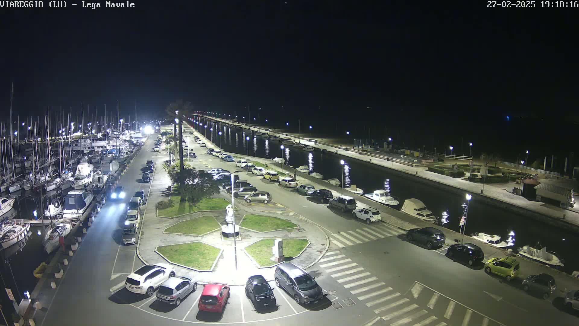 A nighttime view of a marina with numerous boats docked, a paved area with parked cars surrounding a small plaza, and a calm canal reflecting lights.
