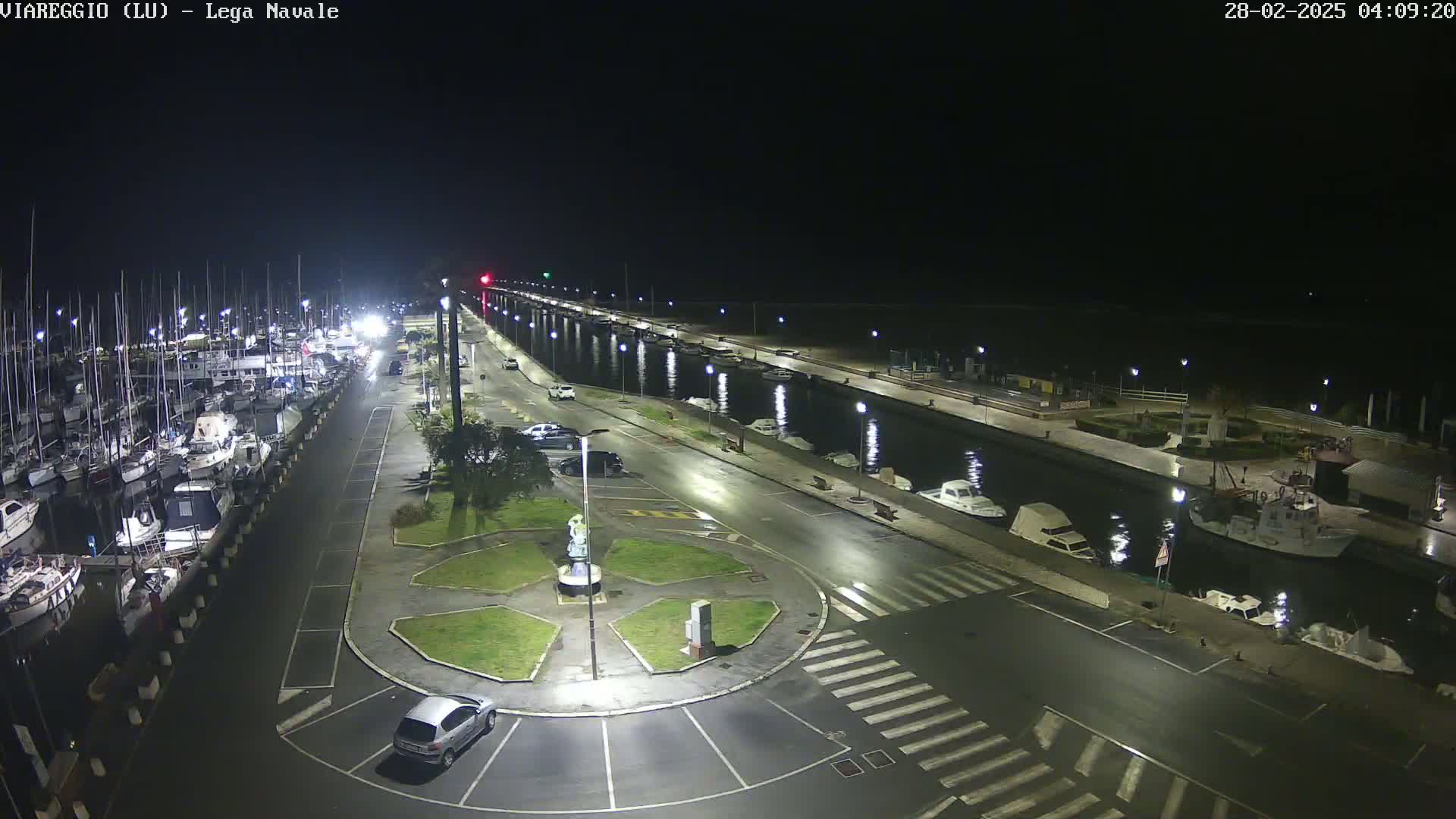 A nighttime view of a harbor filled with boats, a paved road, and a small roundabout with a statue, all under a dark sky.