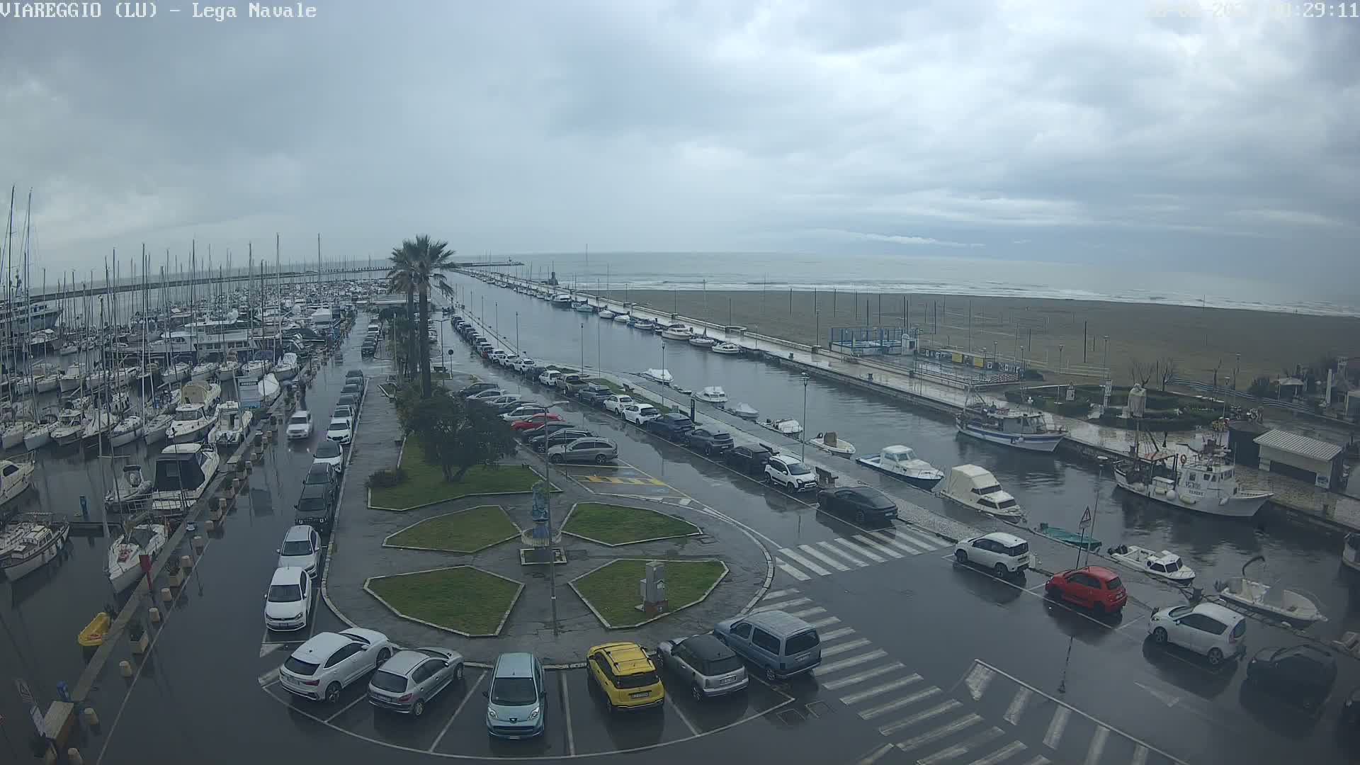 A marina filled with boats is shown, with a flooded parking lot and canal next to a beach under an overcast sky.