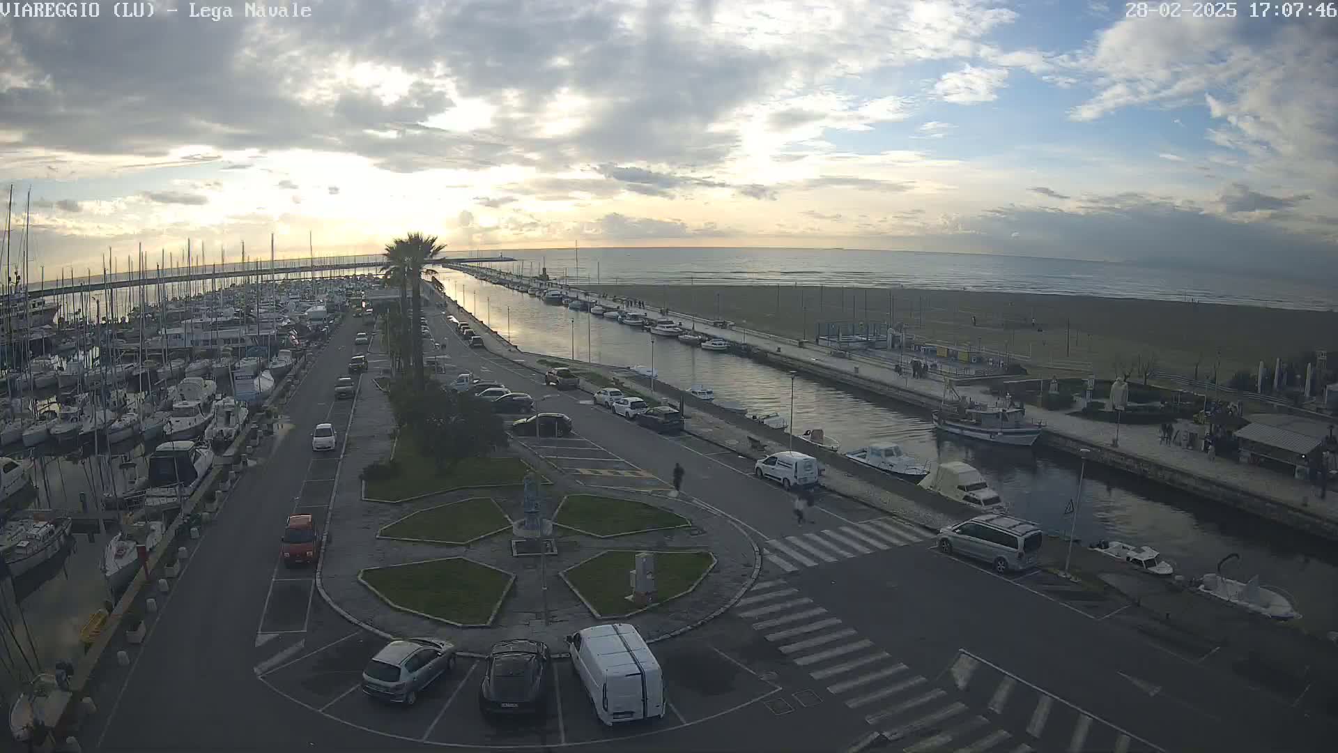 An aerial view shows a marina filled with boats, a canal, a beach, and a parking lot with cars, all under a partly cloudy sky.