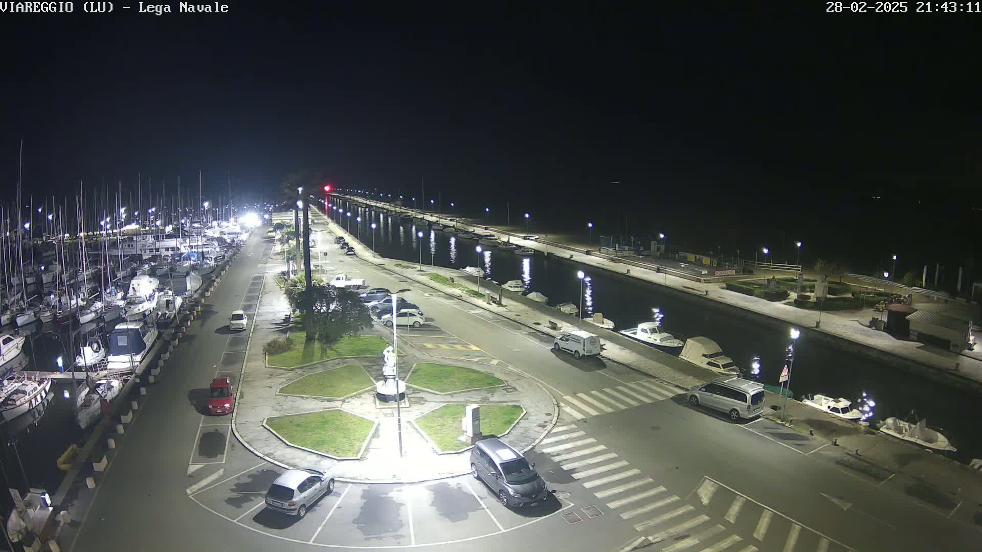 A nighttime aerial view of a marina with docked boats and cars parked along a waterfront road next to a canal.