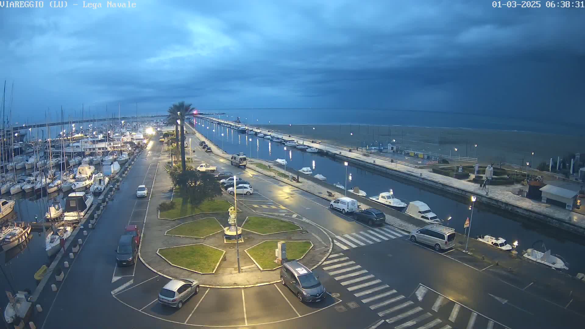 A marina filled with boats at dusk under a cloudy sky, next to a calm sea and a roadway with parked cars.