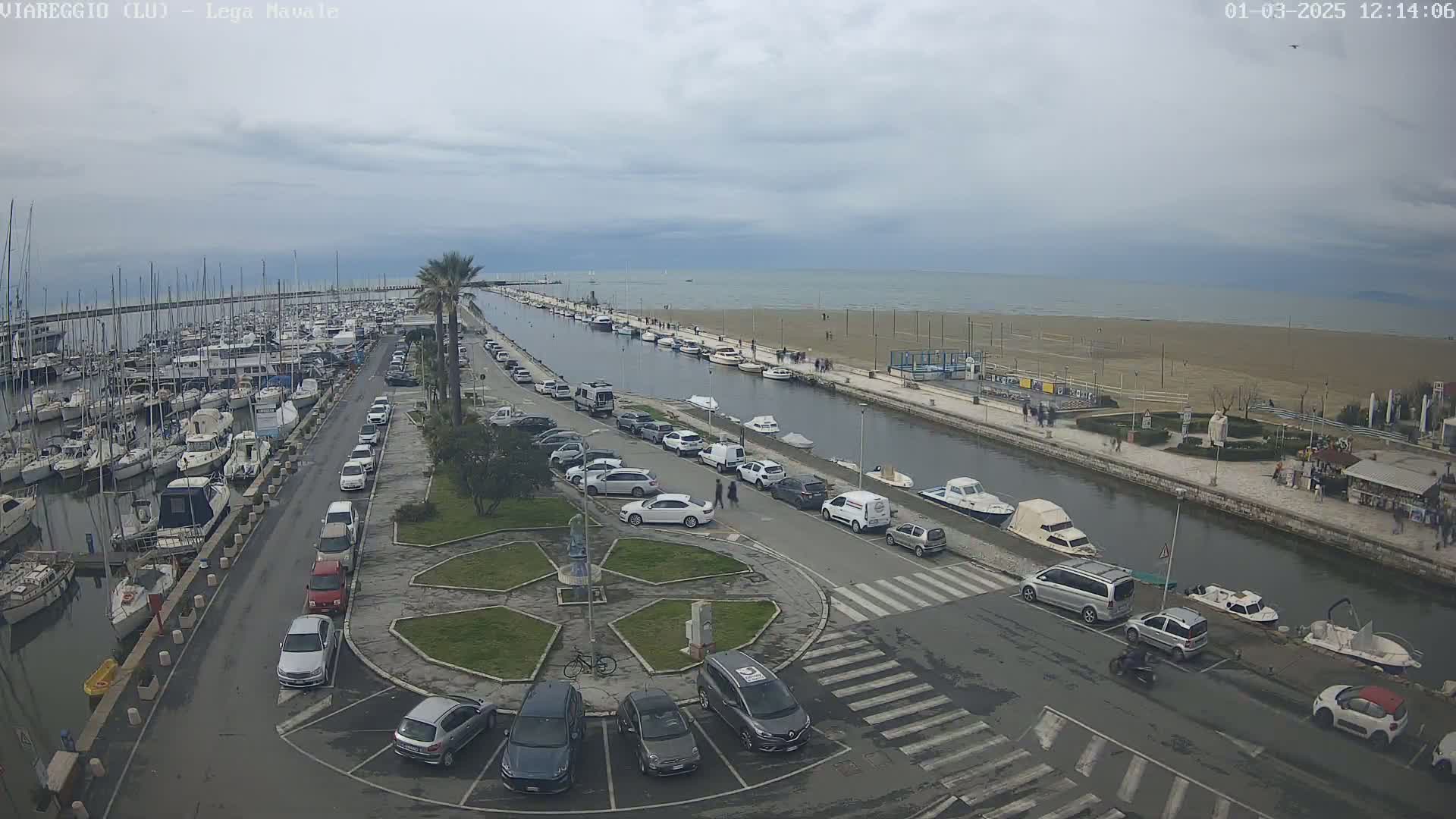 An overcast day at a harbor shows a marina filled with boats, a canal with moored boats, cars parked along the waterfront, and a sandy beach beyond.