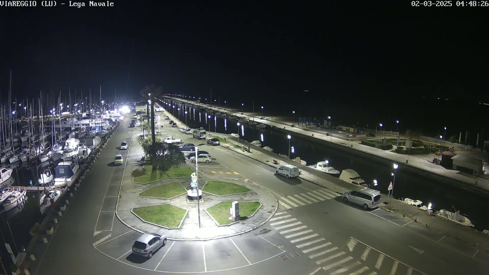 A nighttime aerial view of a marina with numerous boats docked, a roadway with parked cars, and a small roundabout in the center, all illuminated by streetlights.