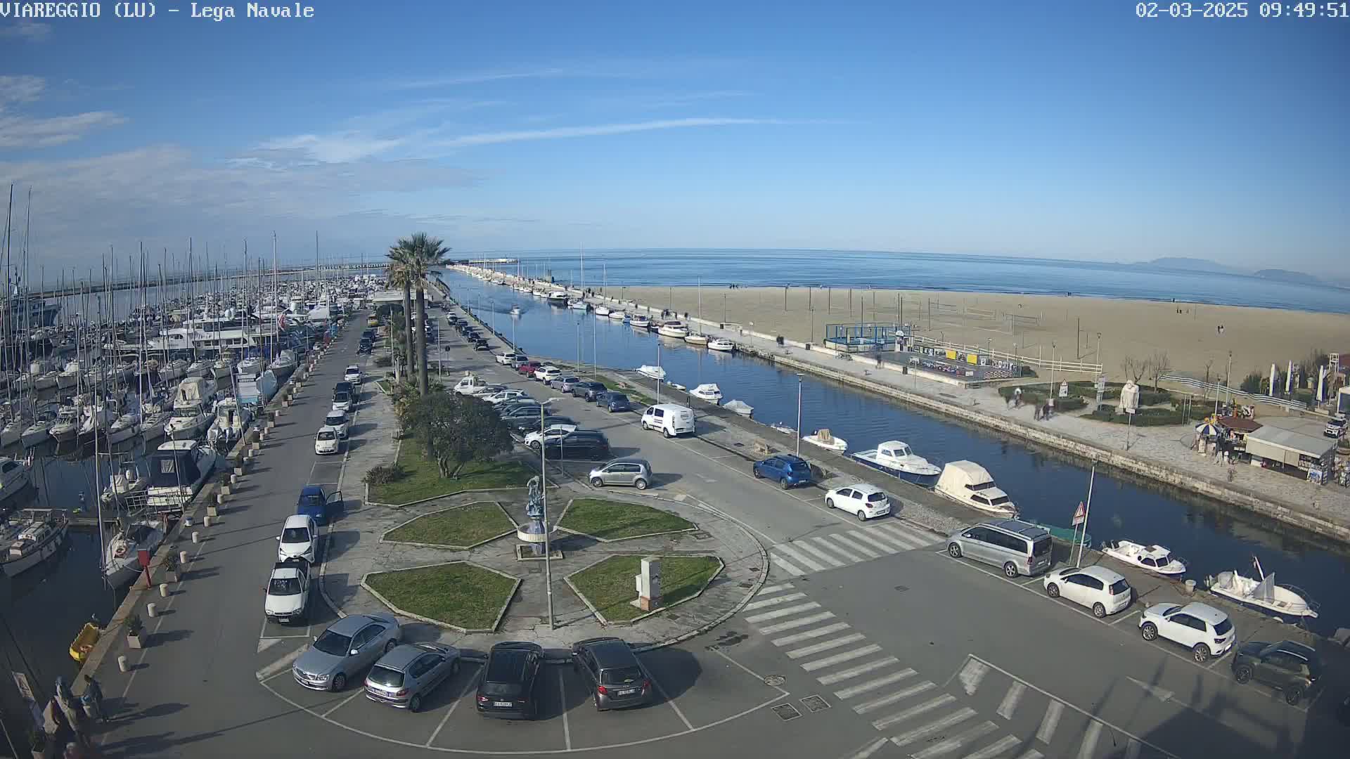 A marina filled with boats is adjacent to a sandy beach and calm ocean under a clear blue sky.