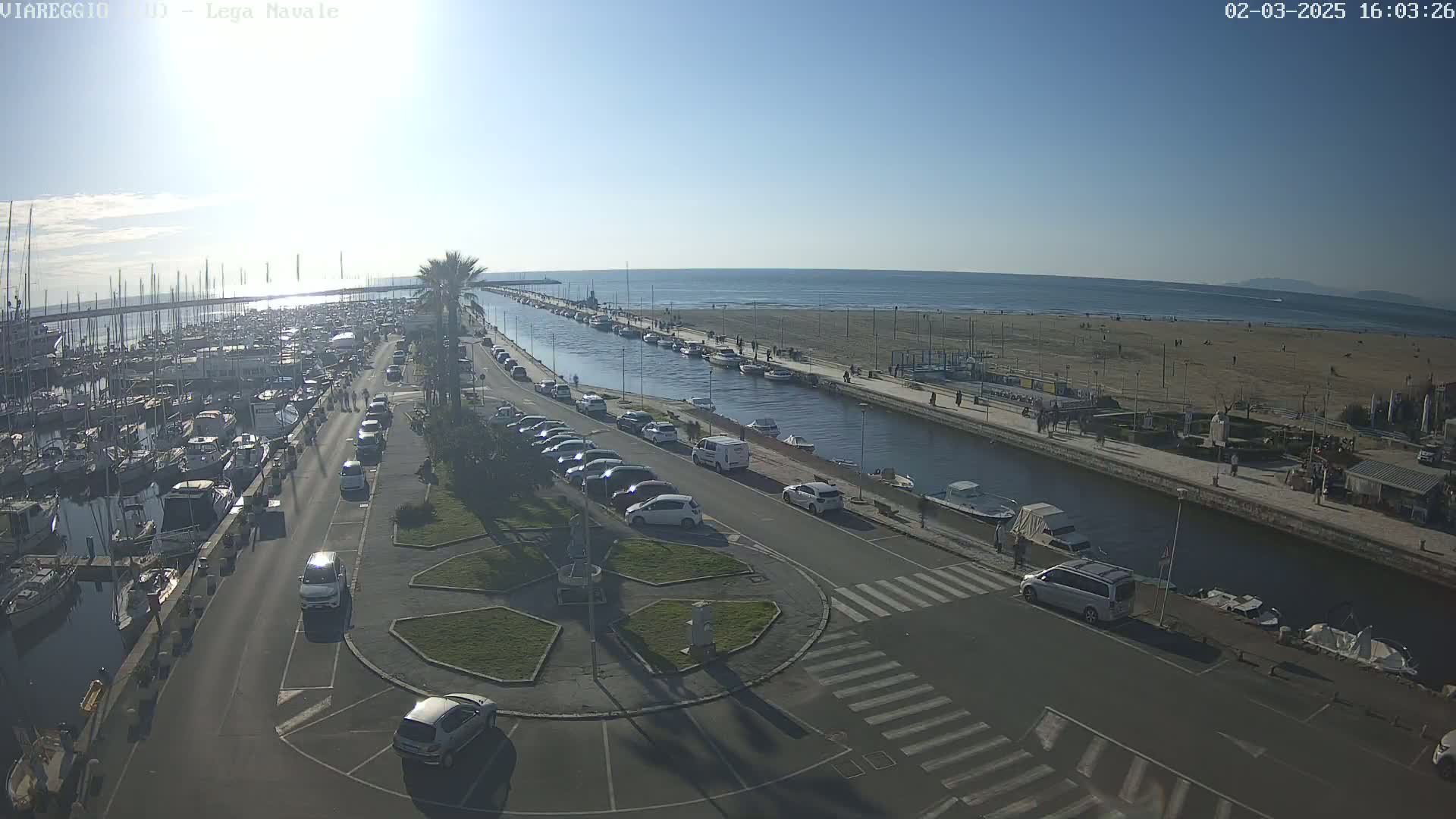 A sunny day shows a harbor filled with boats, a canal with parked cars along its sides, and a sandy beach beyond.