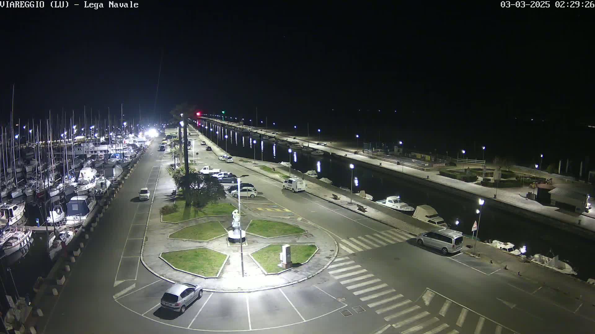 A nighttime aerial view of a marina with numerous boats docked, a roadway with parked cars, and a circular landscaped area.