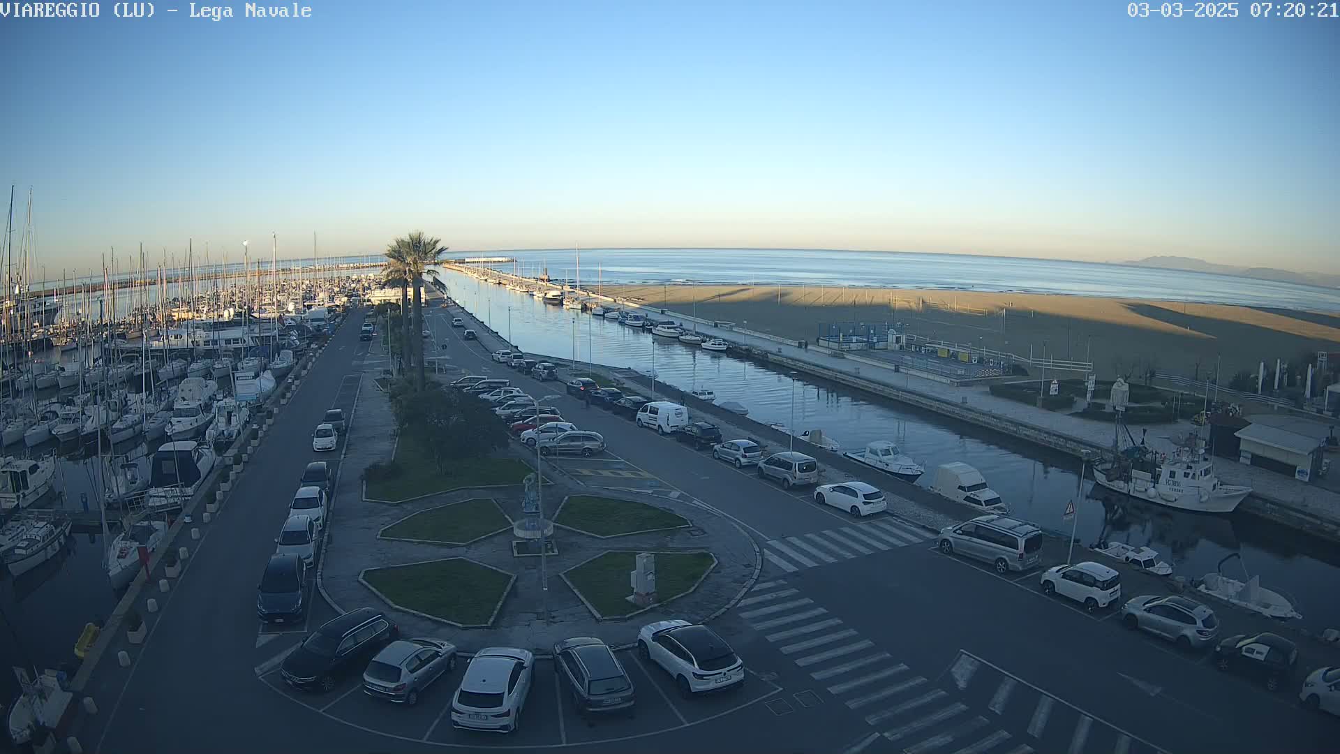 A marina filled with boats is next to a canal that leads to a sandy beach and calm ocean under a clear blue sky.