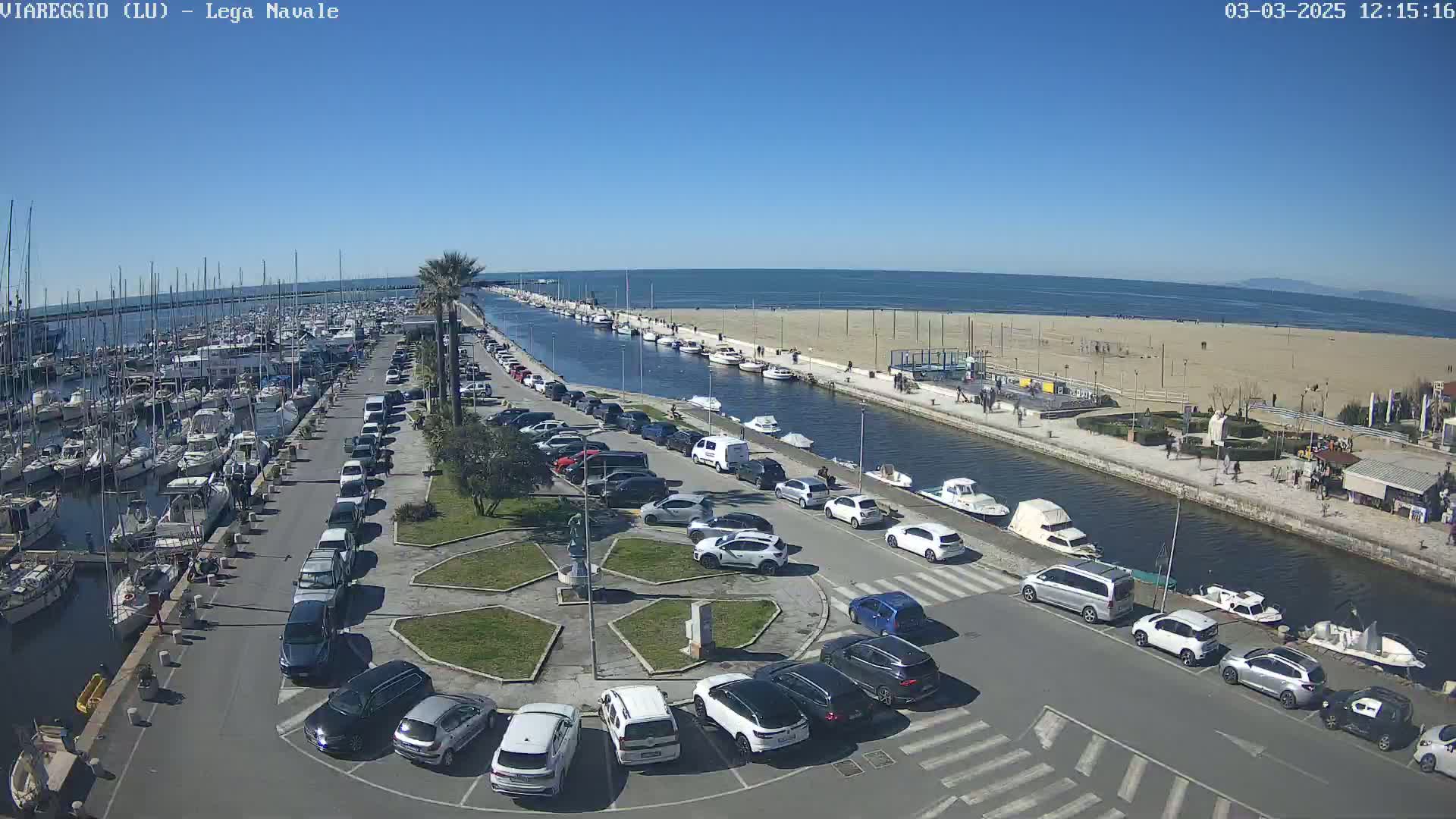 A marina filled with boats is next to a sandy beach and a canal lined with parked cars under a clear blue sky.