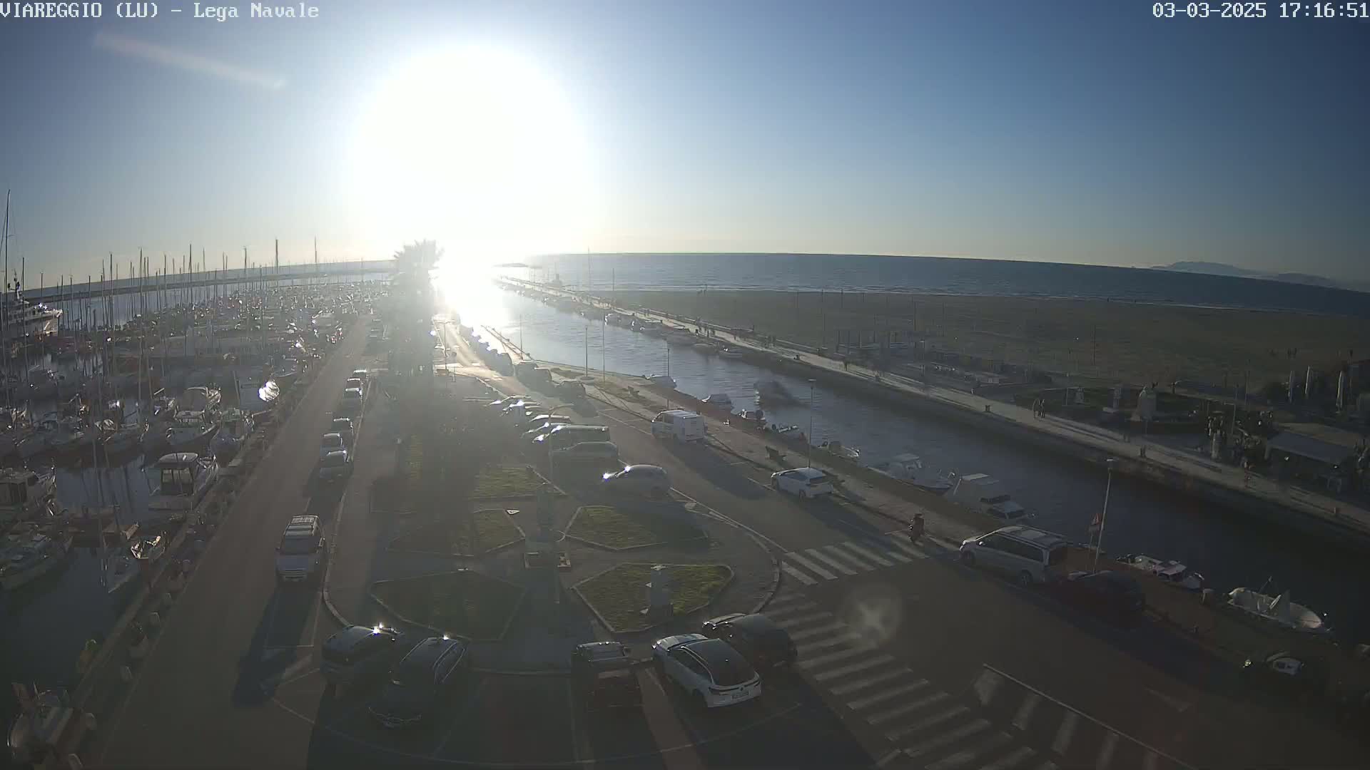 A sunny harbor scene with a marina full of boats, a canal, a beach, and cars parked along a road.