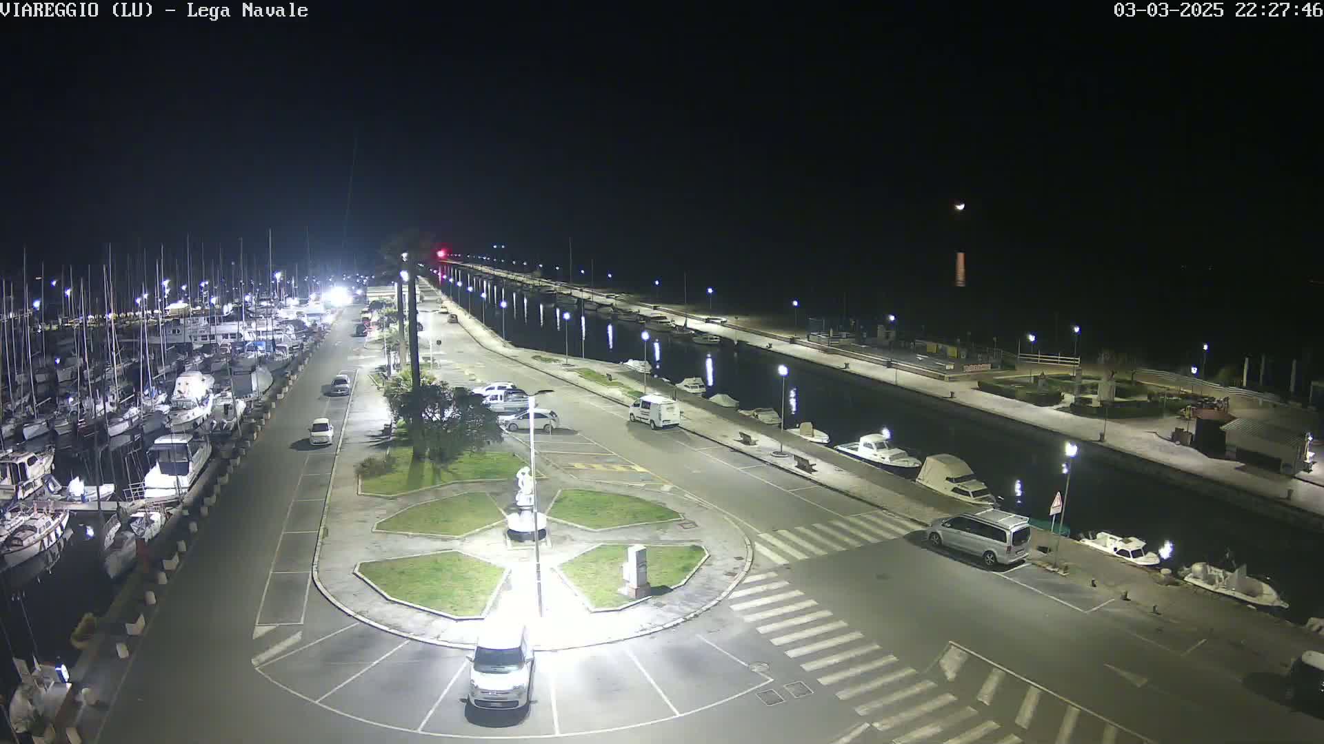 A nighttime view of a marina with numerous boats docked, a paved area with cars parked, and a small roundabout in the center, all brightly lit under a clear, dark sky.